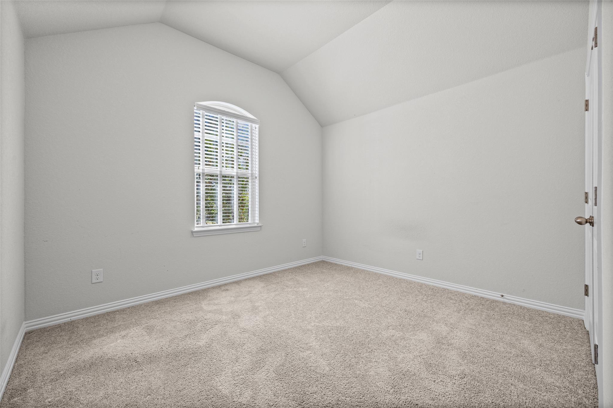 Empty carpeted room with a vaulted ceiling and an arched window with white shutters.