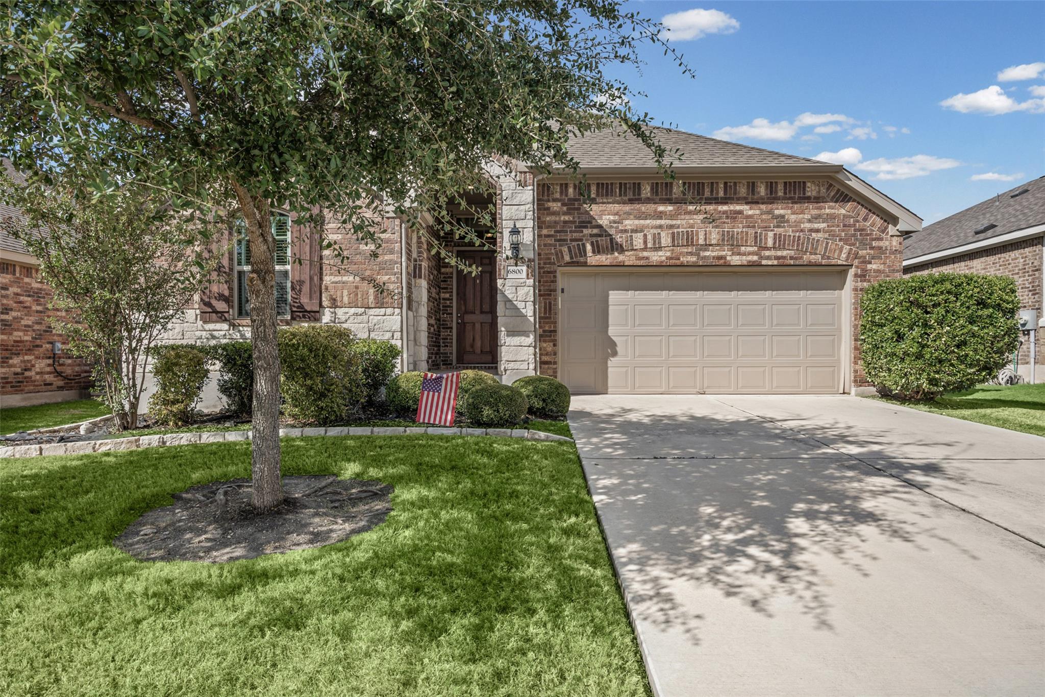 Brick and stone home with a two-car garage, mature front-yard tree, and American flag.