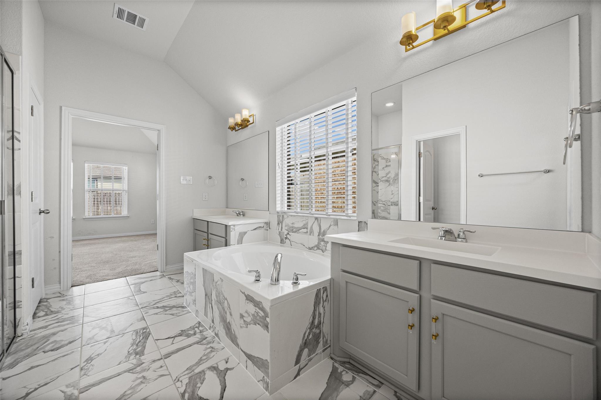 Bathroom with a marble-clad soaking tub, gray double vanity, and large window with blinds.