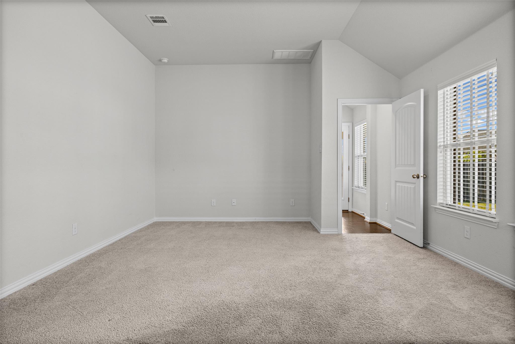 Empty carpeted room with a high angled ceiling, a window with blinds, and an open door to the hall.