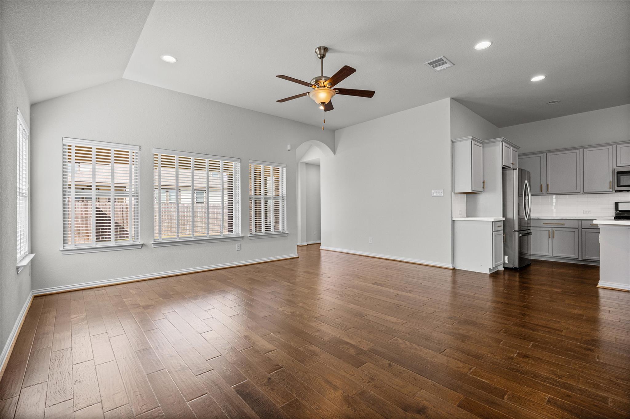 Open living and dining area with brown flooring, large windows, ceiling fan, and adjoining kitchen.