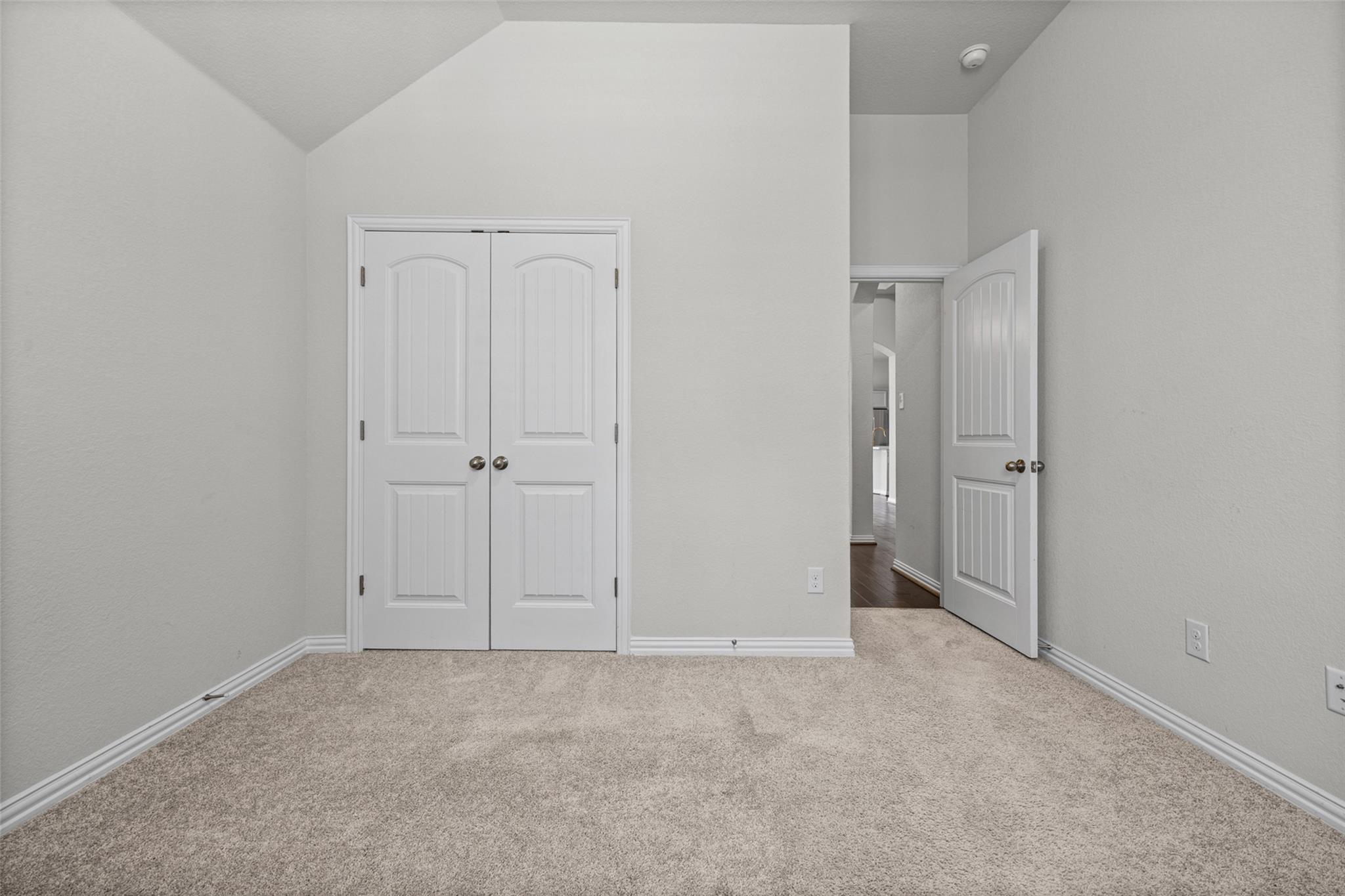 Empty carpeted bedroom with a vaulted ceiling, double closet doors, and an open hallway doorway.