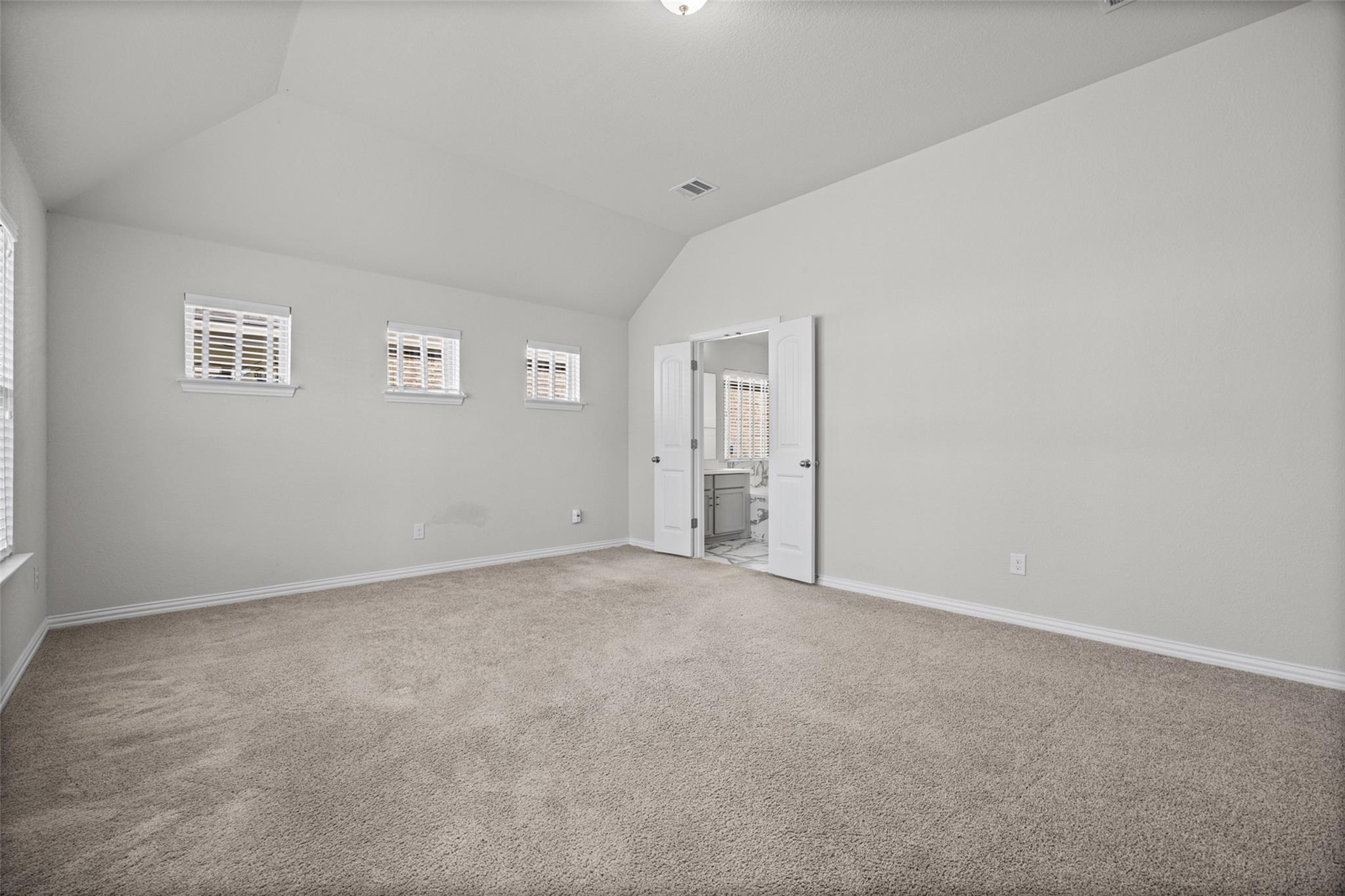 Empty carpeted bedroom with a vaulted ceiling, three small windows, and open doors to a bathroom.