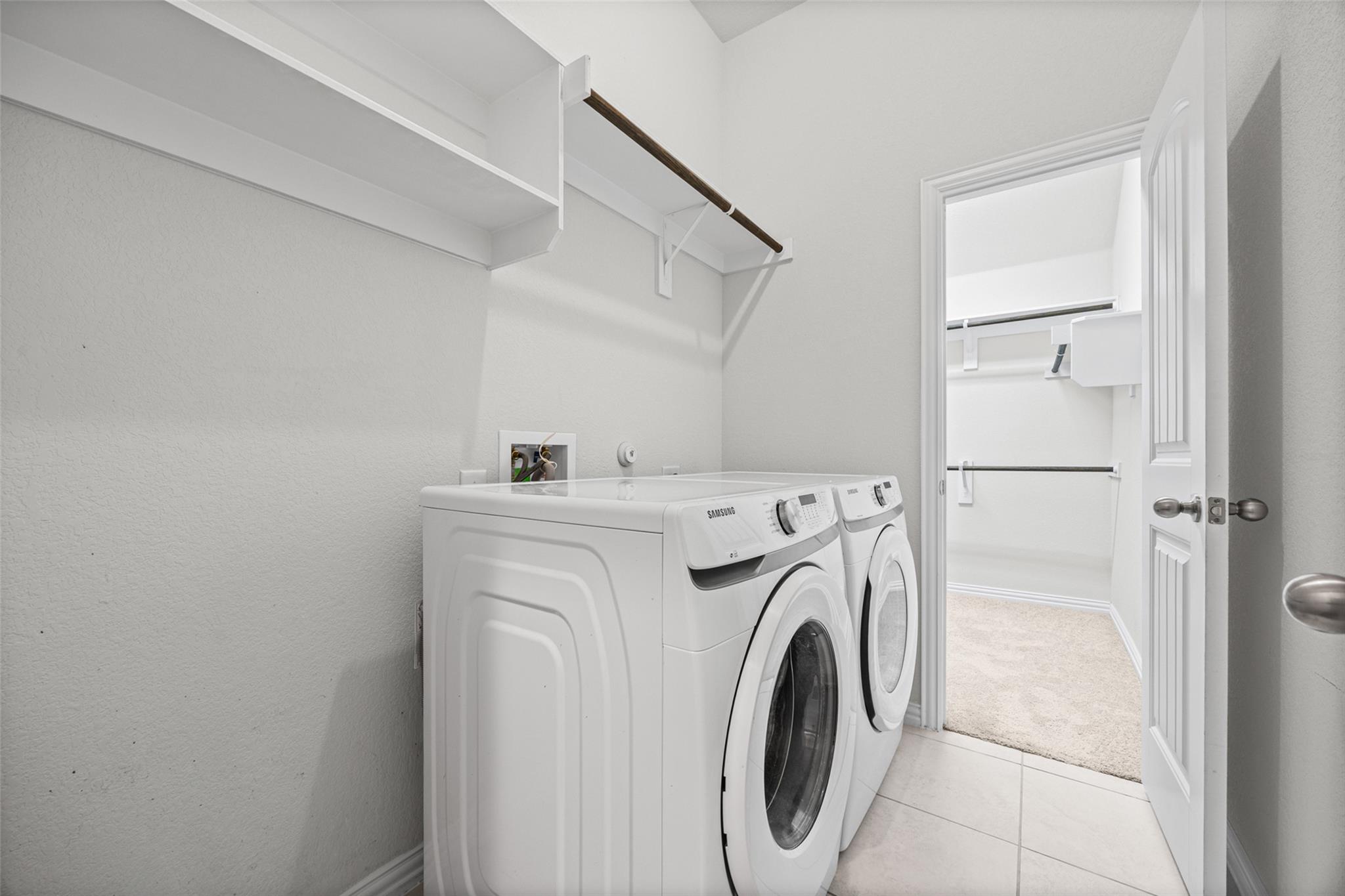 Laundry room with white front-loading washer and dryer, overhead shelves, and an open doorway to a carpeted closet.