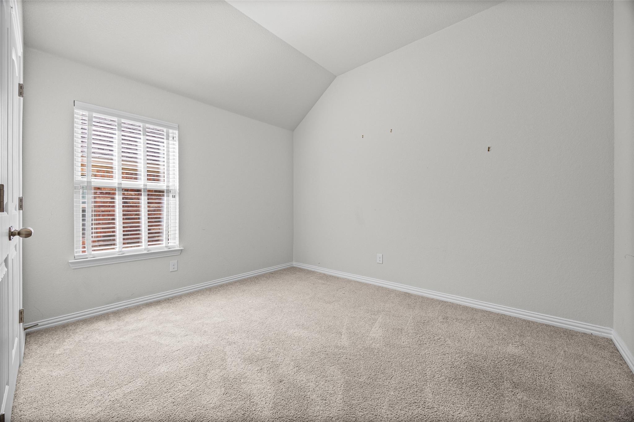 Empty bedroom with beige carpet, a high angled ceiling, and a shuttered window.