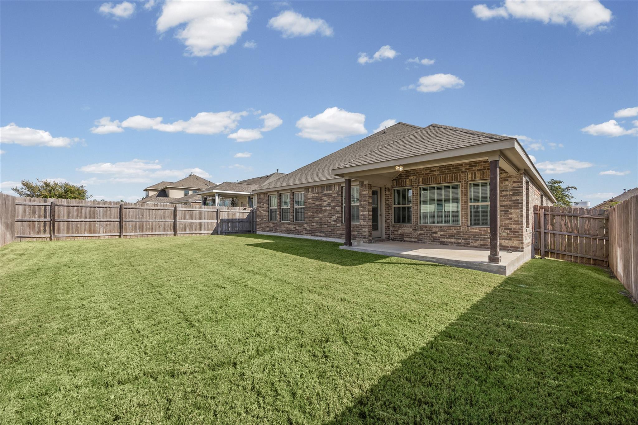 Brick home's covered back patio overlooks a spacious green lawn enclosed by wooden fencing.