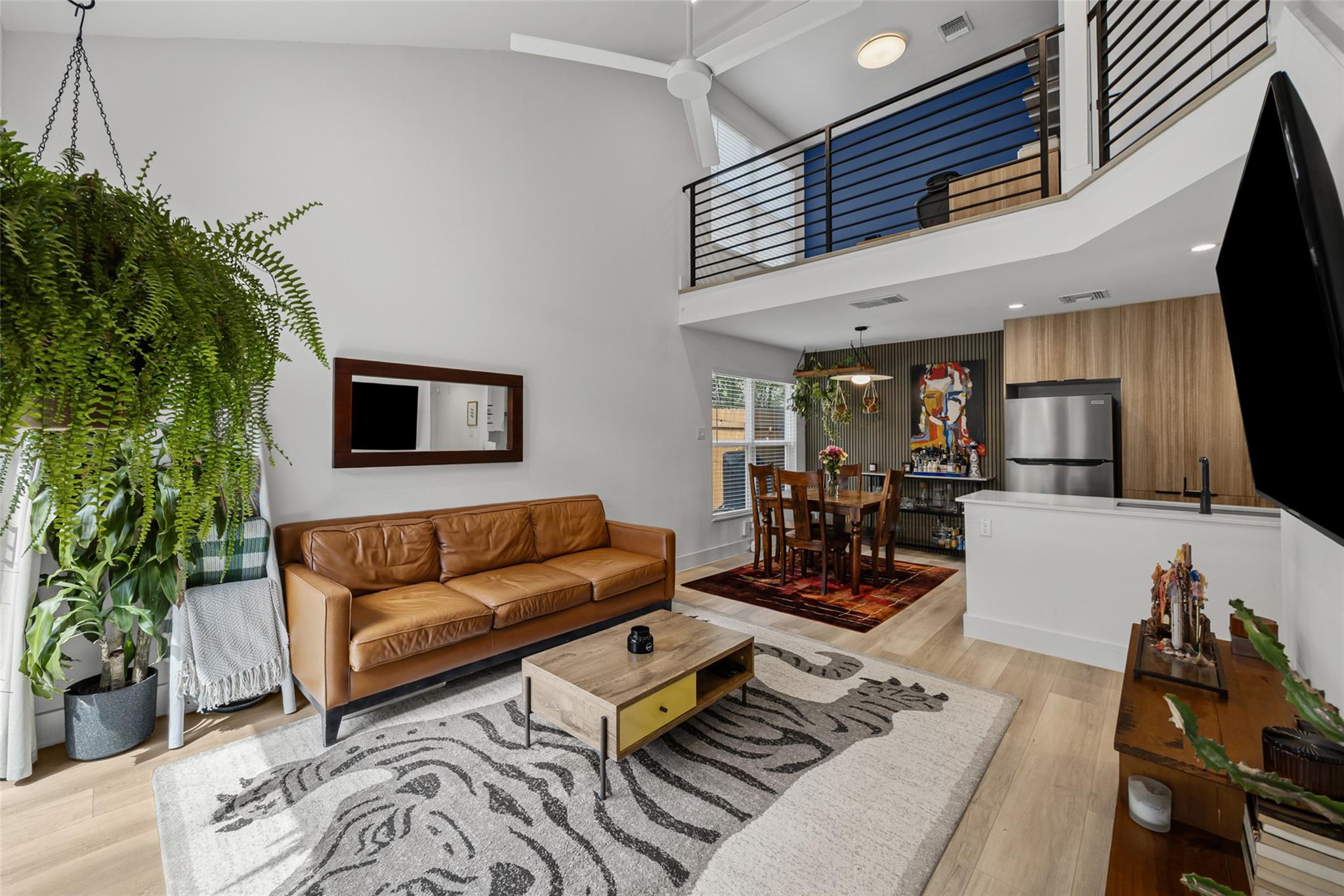 Open-plan living room with a leather sofa, hanging fern, kitchen island, and upstairs balcony.