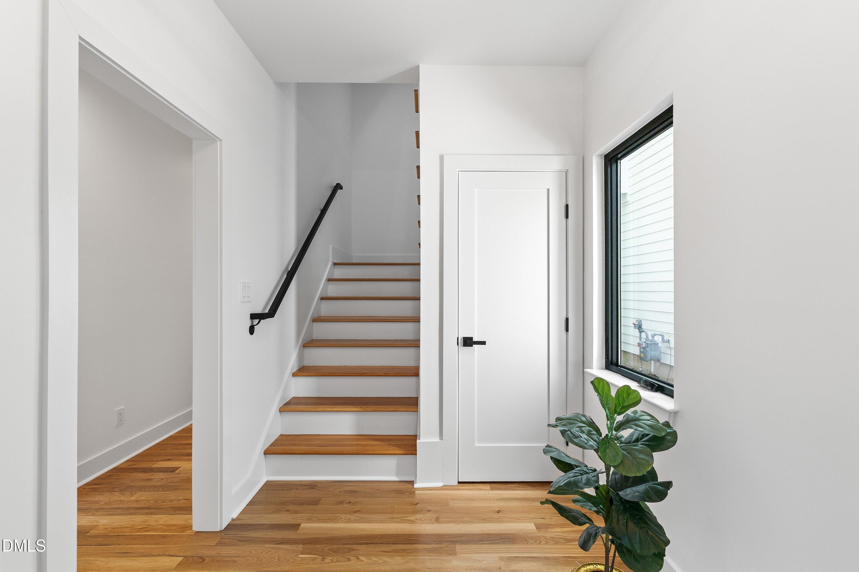 Entryway with wood staircase, black handrail, hardwood floors, and a leafy plant by the window.