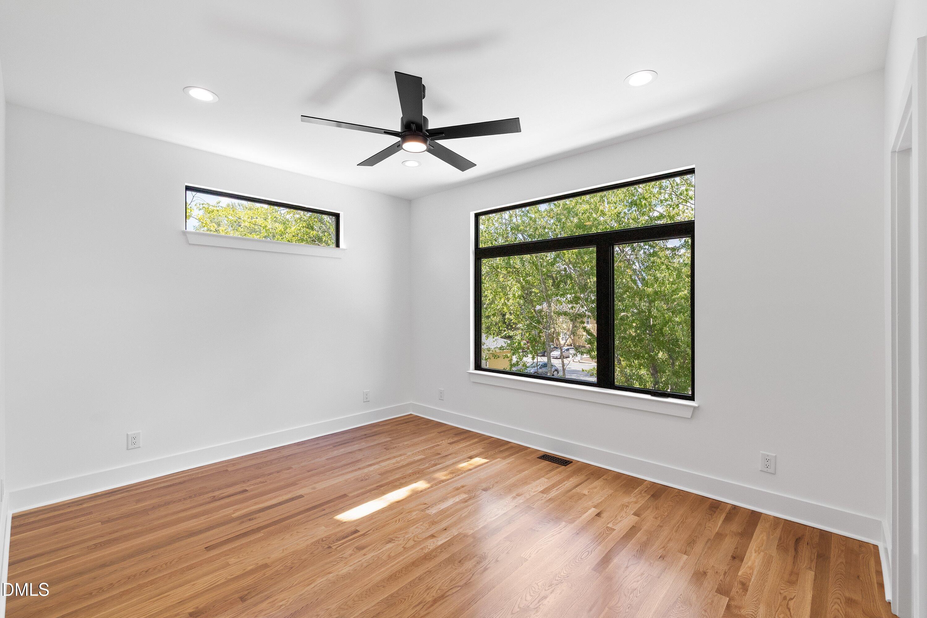 Empty room with hardwood floors, a black ceiling fan, and large windows overlooking leafy trees.