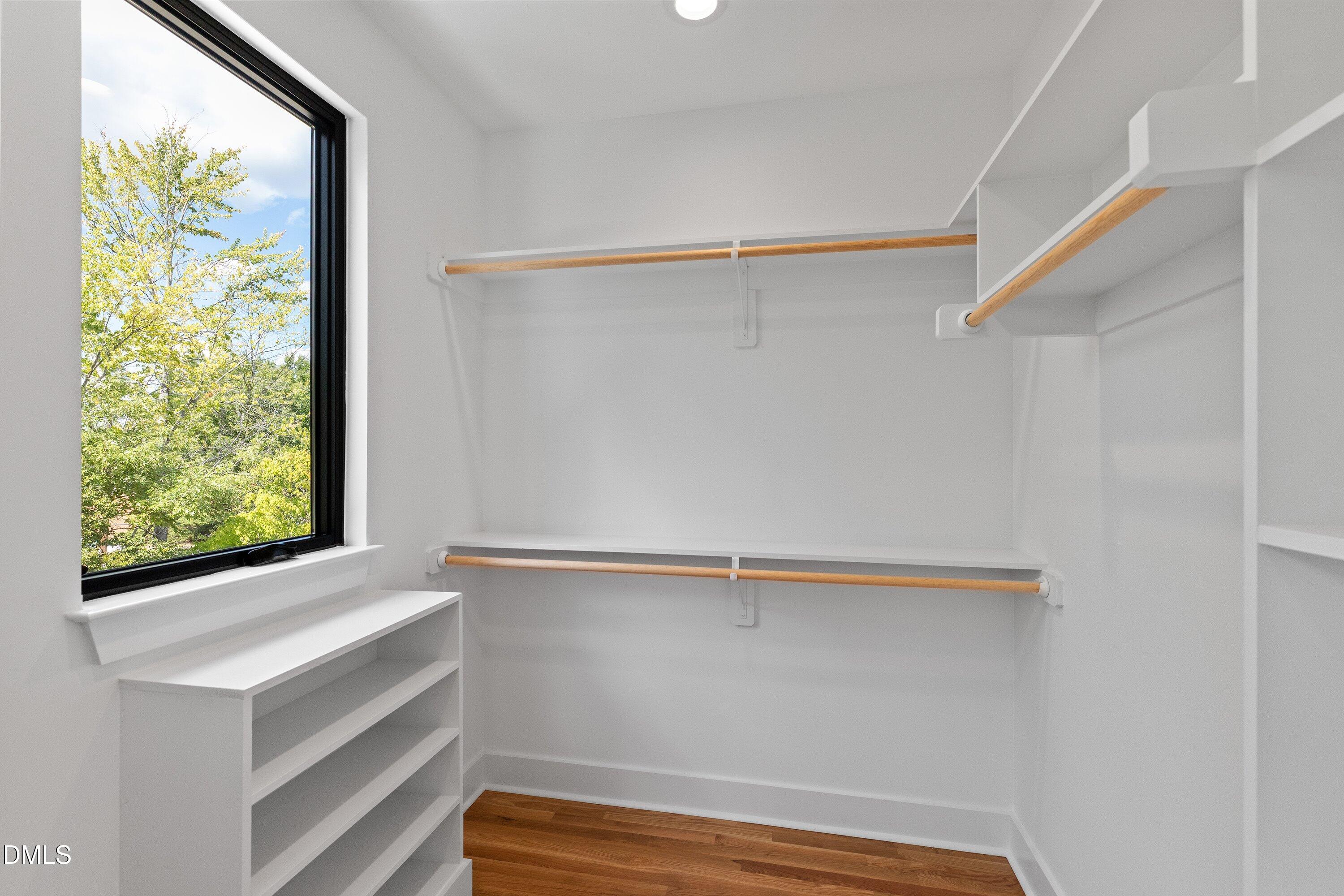 Walk-in closet with built-in shelves, wooden hanging rods, hardwood floors, and a large window.
