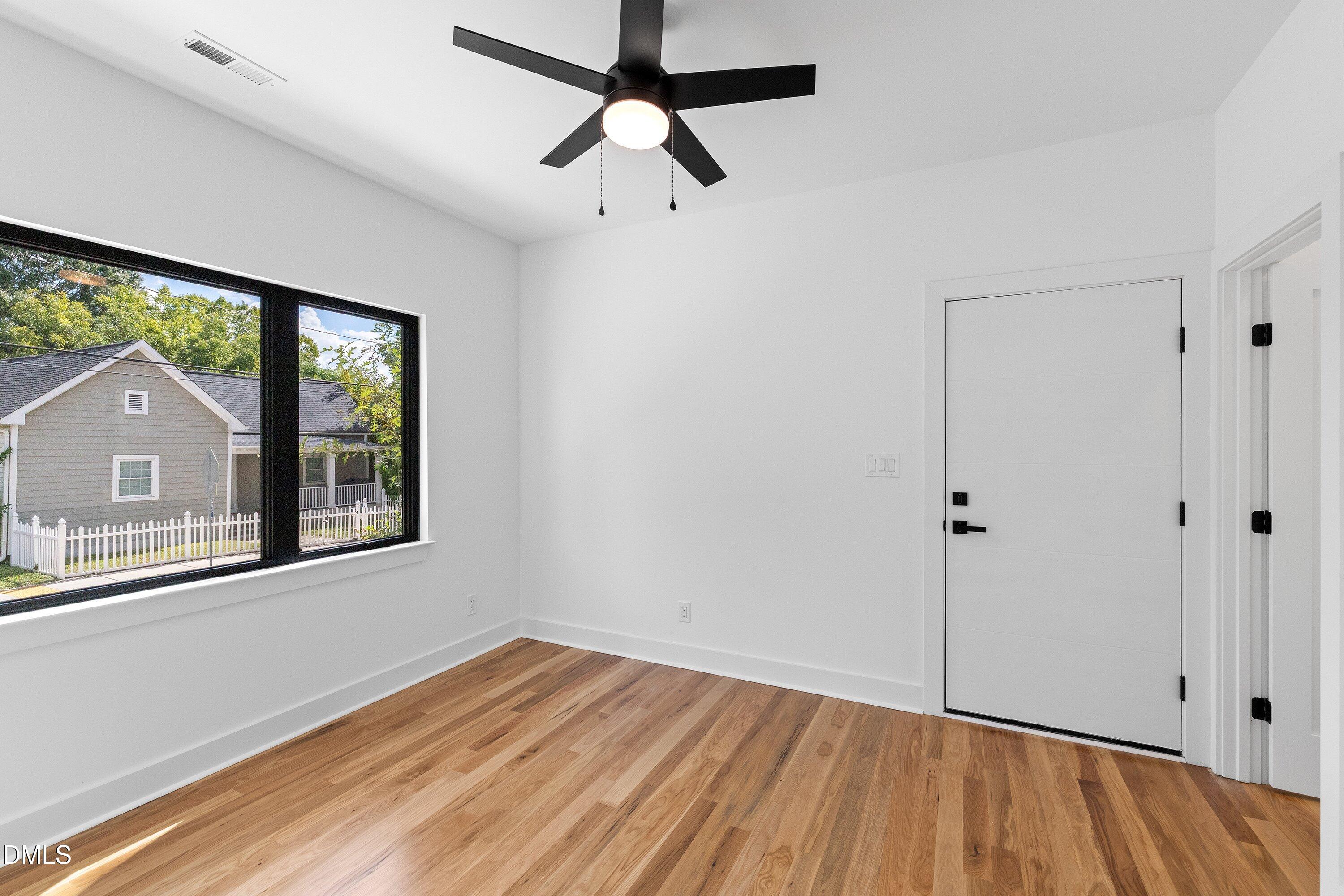 Empty room with hardwood floors, a black ceiling fan, and a large window overlooking a neighboring home.