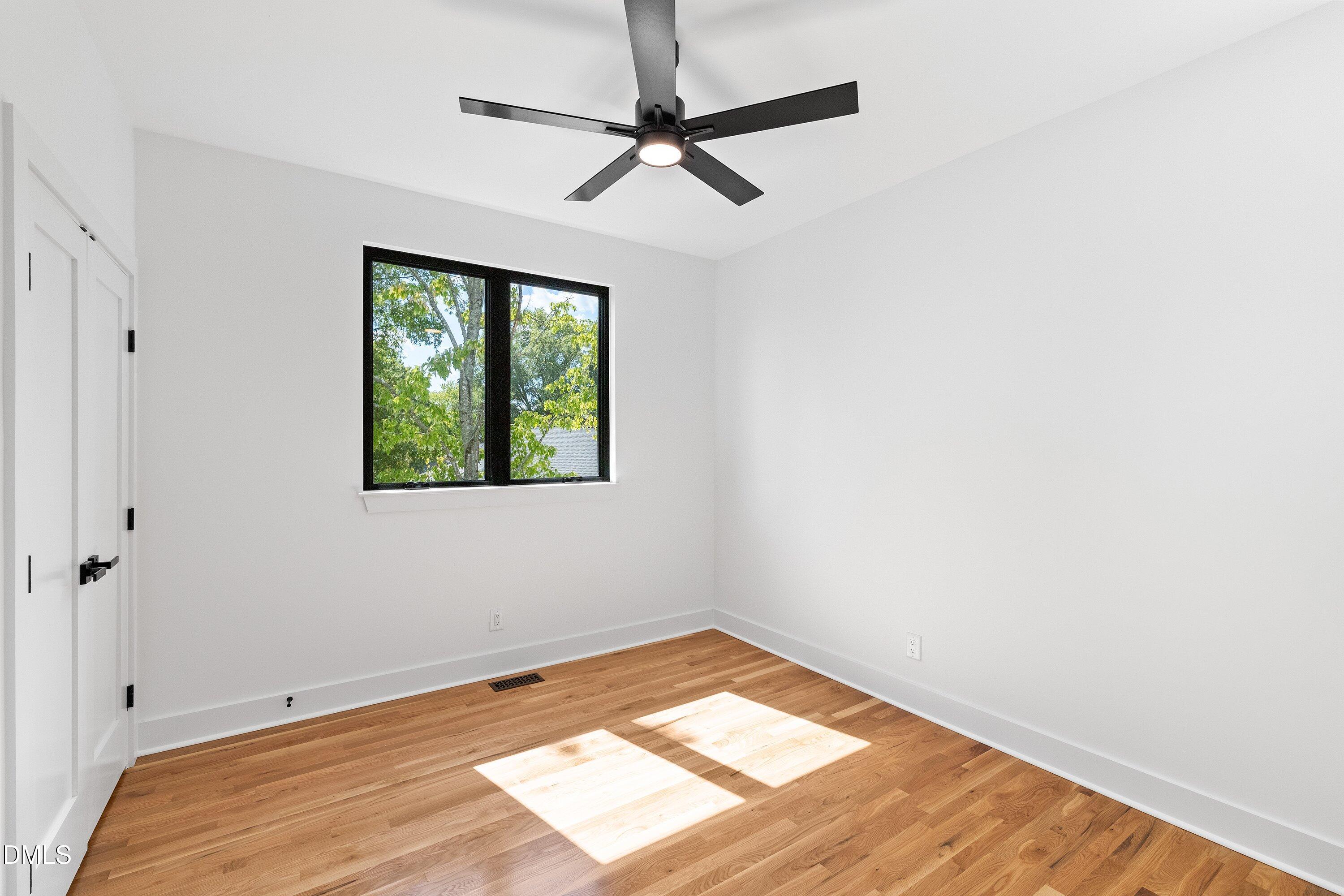 Empty bedroom with hardwood floors, a black ceiling fan, and sunlight streaming through a window.