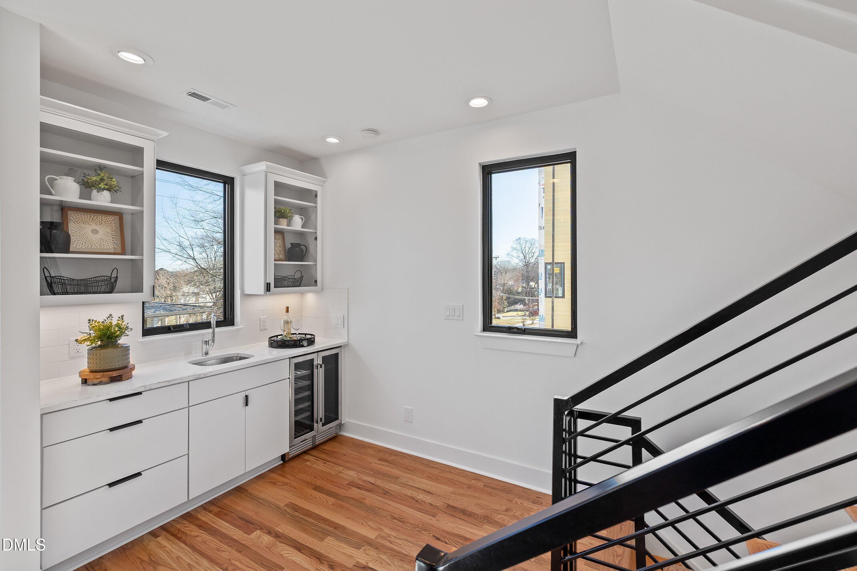 Kitchenette with white cabinets and open shelves beside a black metal staircase and warm wood floors.