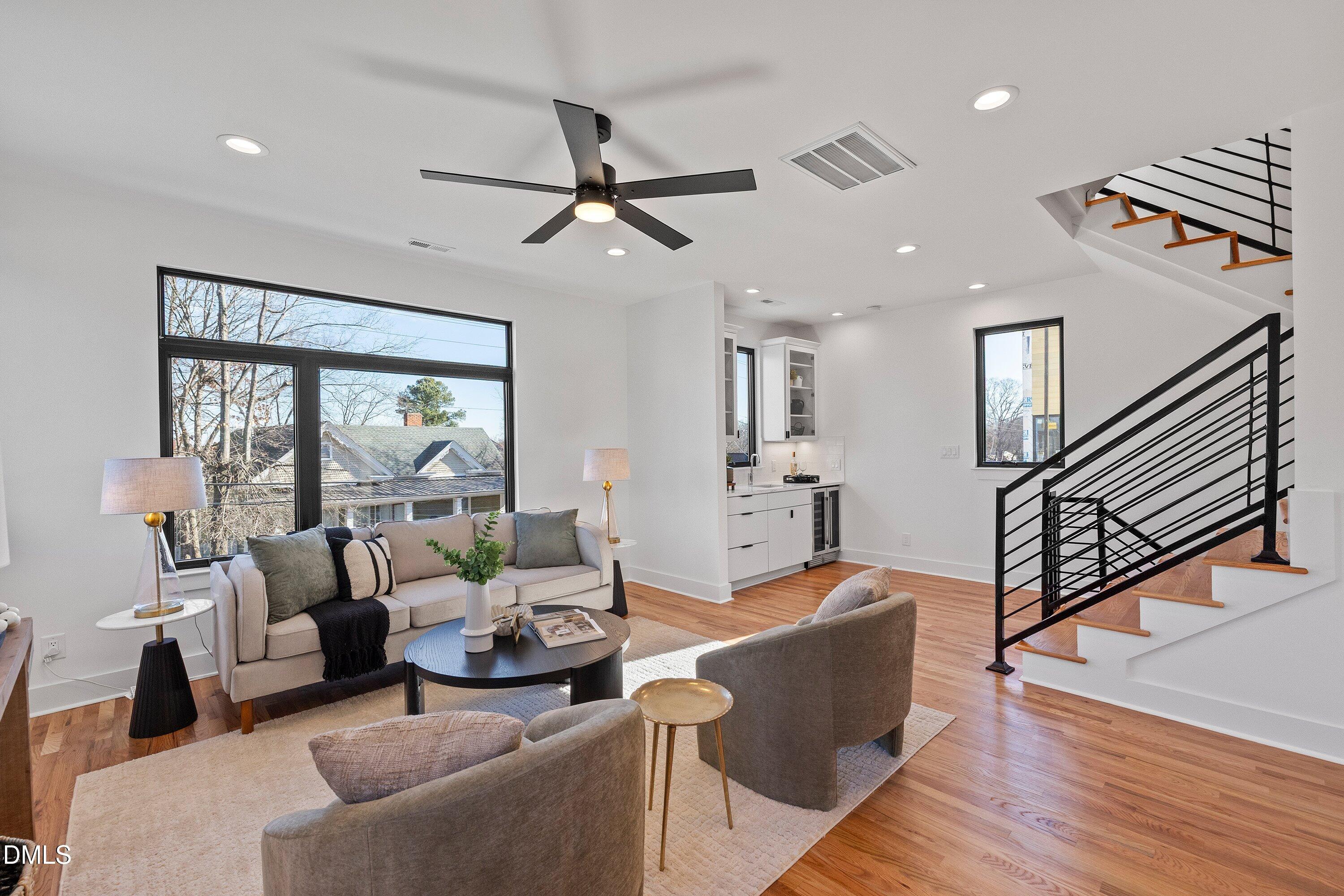 Staged living room with large windows, light wood floors, an open kitchen, and a black-railed staircase.