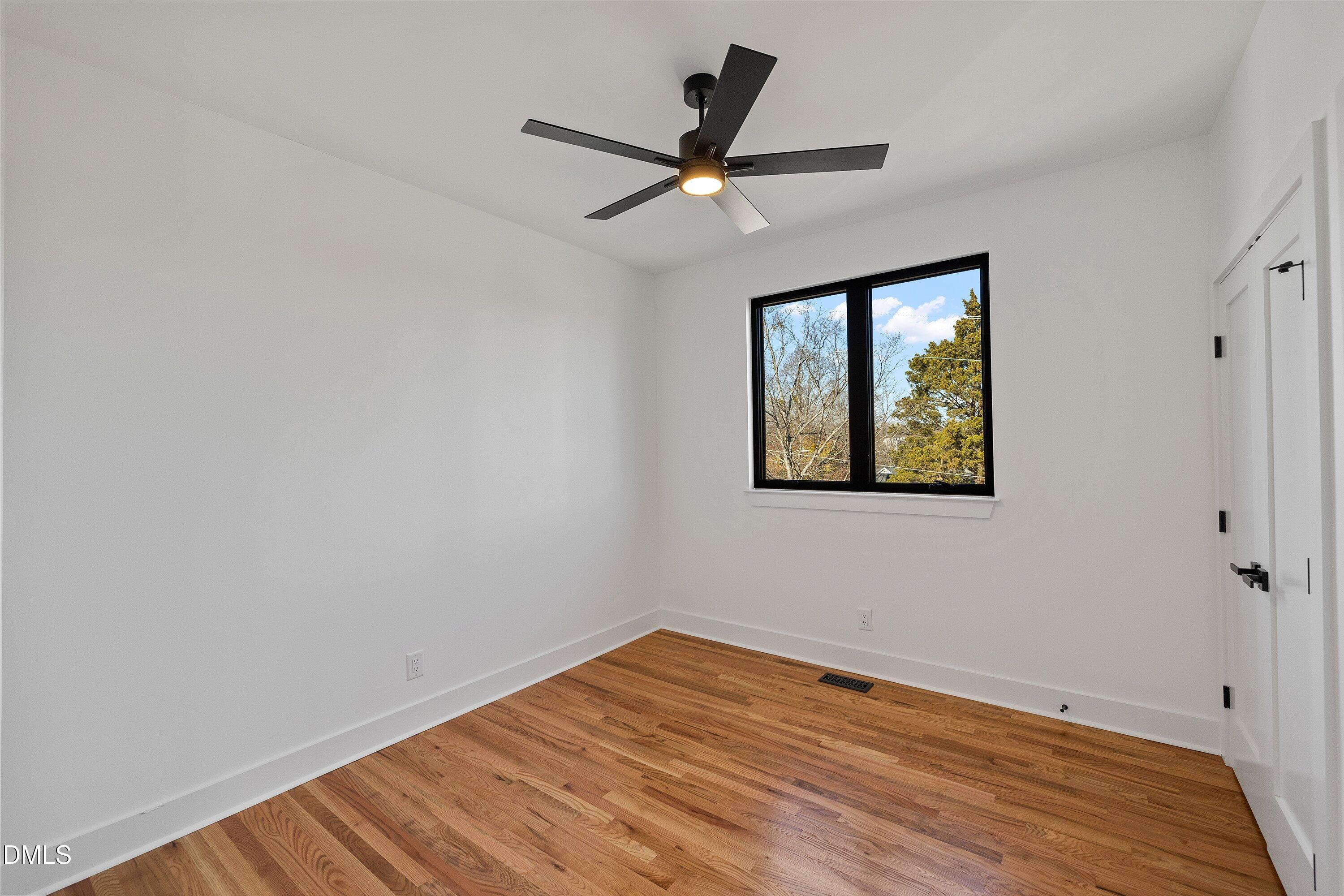 Vacant bedroom with hardwood floors, a black ceiling fan, and a window overlooking trees.