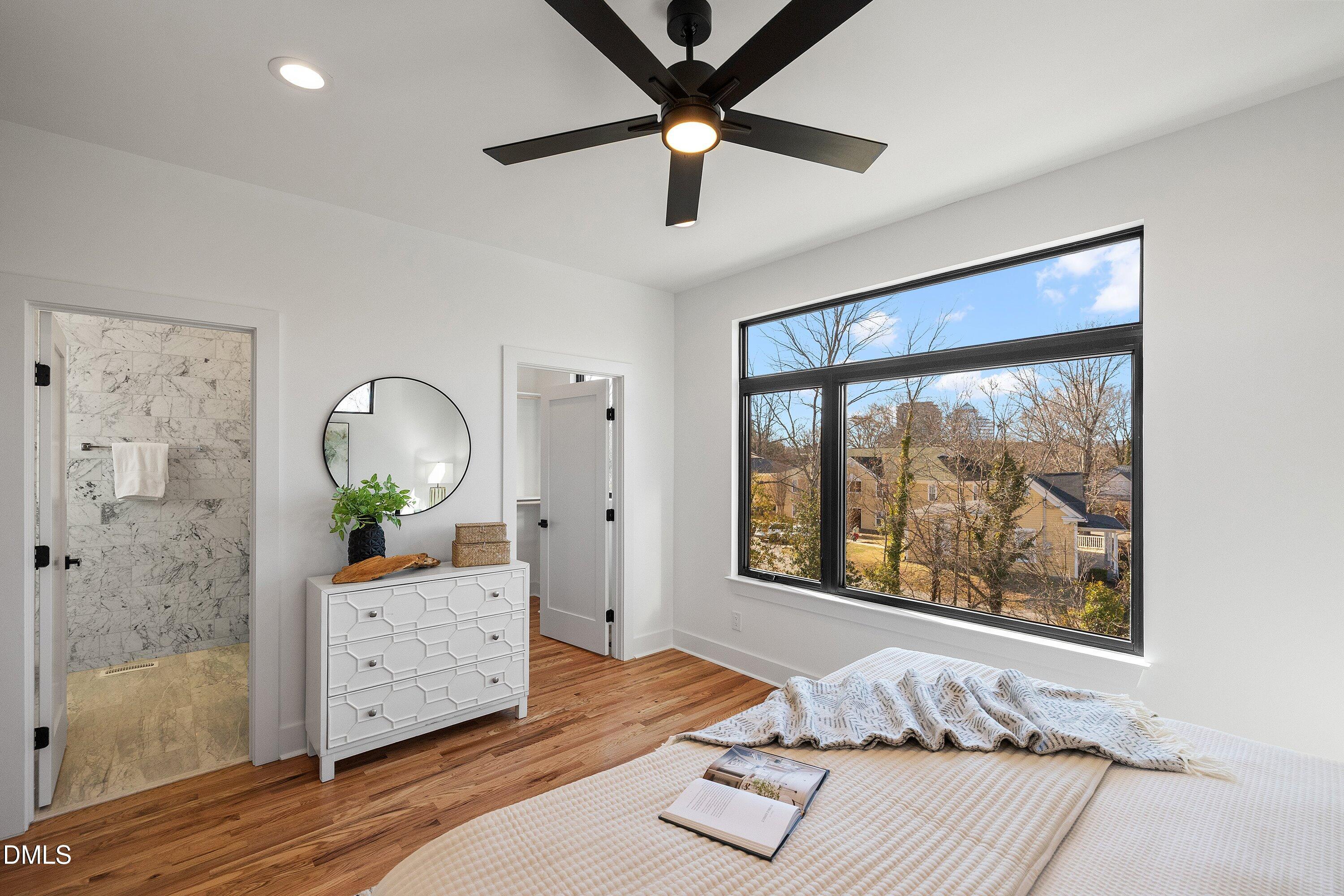 Staged bedroom with hardwood floors, a large neighborhood-view window, and an adjoining marble-tiled shower.