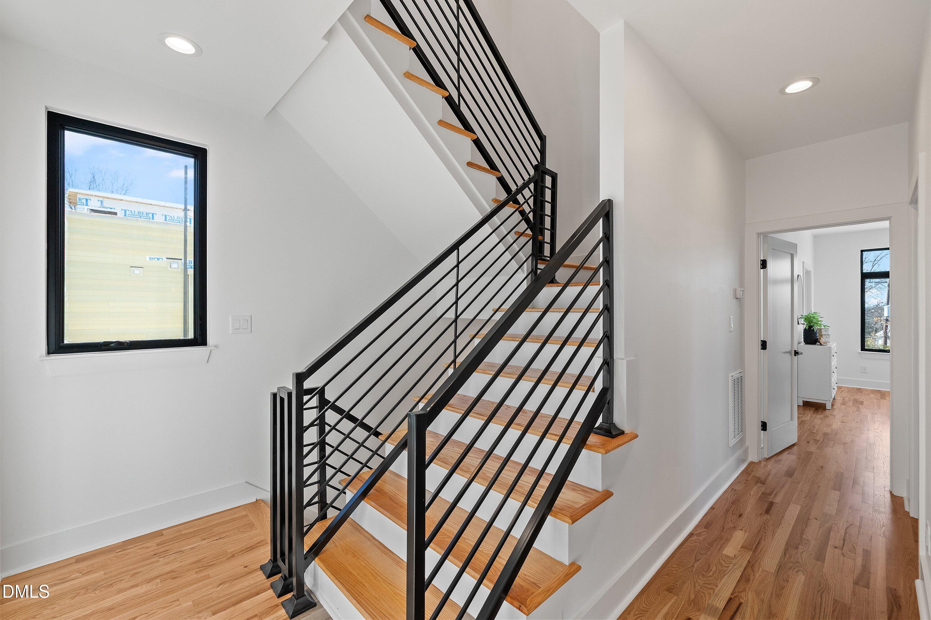 Open wood staircase with black metal railings beside a large window and hardwood-floored hallway.