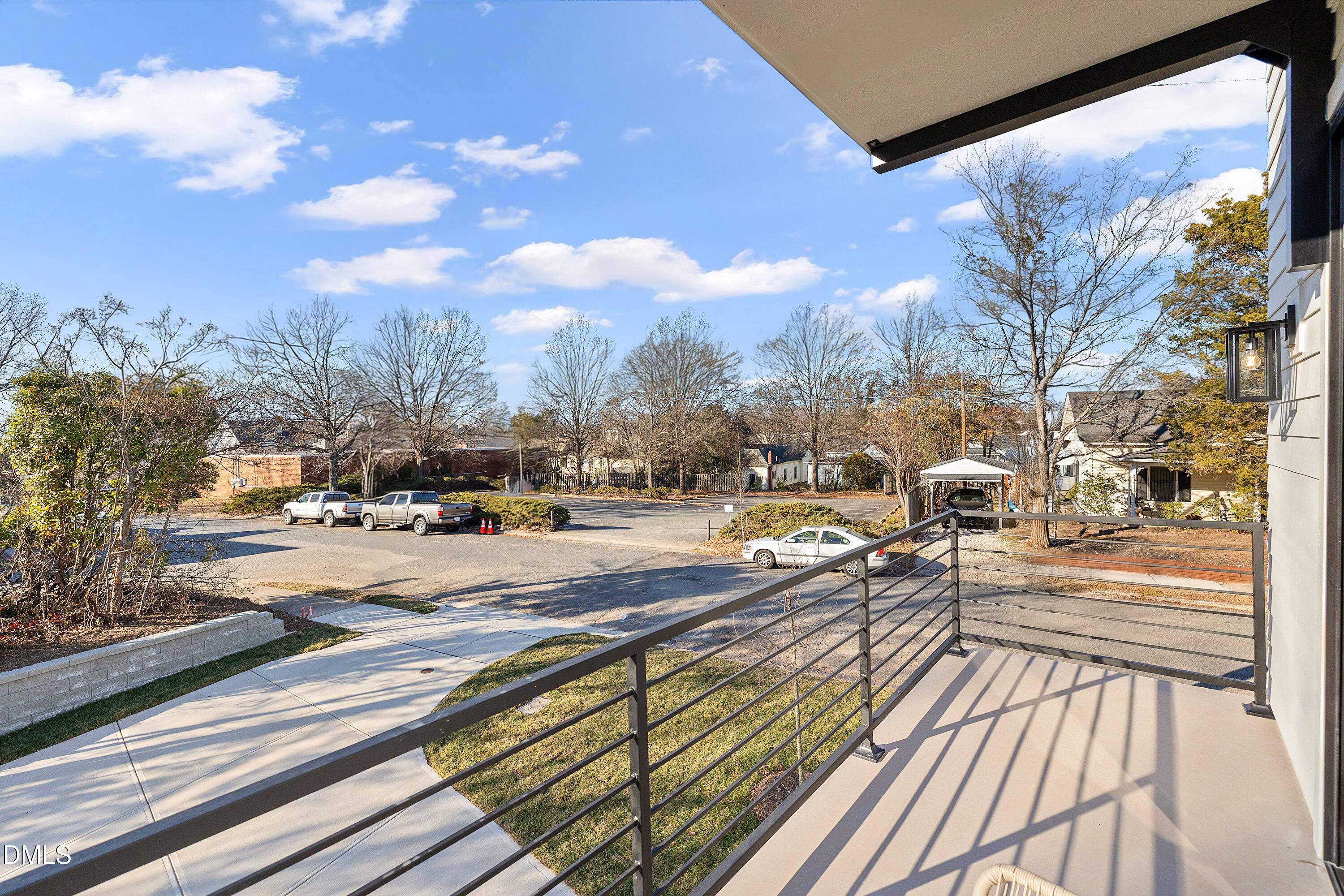 Covered balcony with dark railings overlooking a sidewalk, parking area, and neighboring buildings.