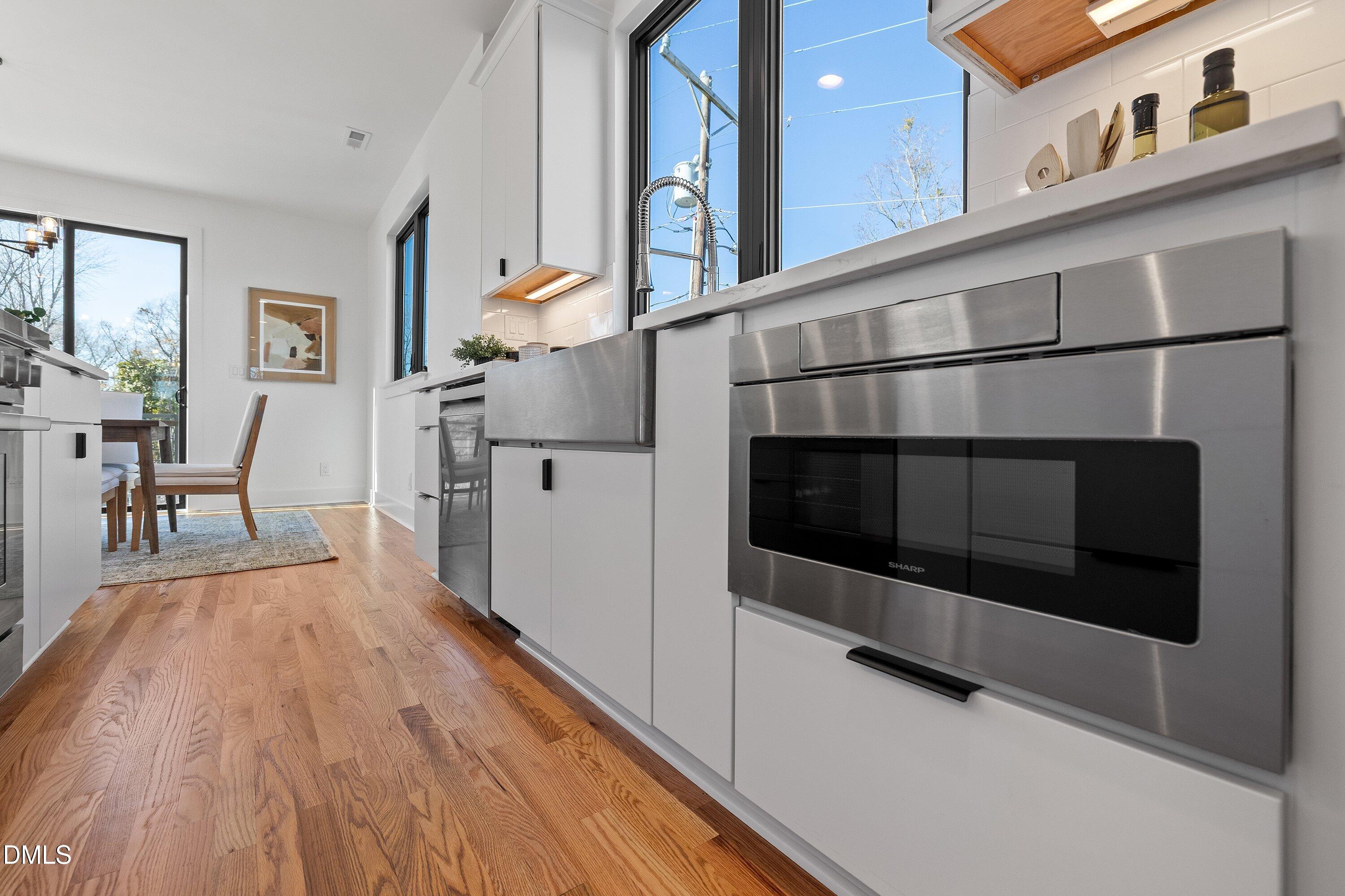 Modern white kitchen with stainless steel appliances, hardwood floors, and windows beside a small dining area.