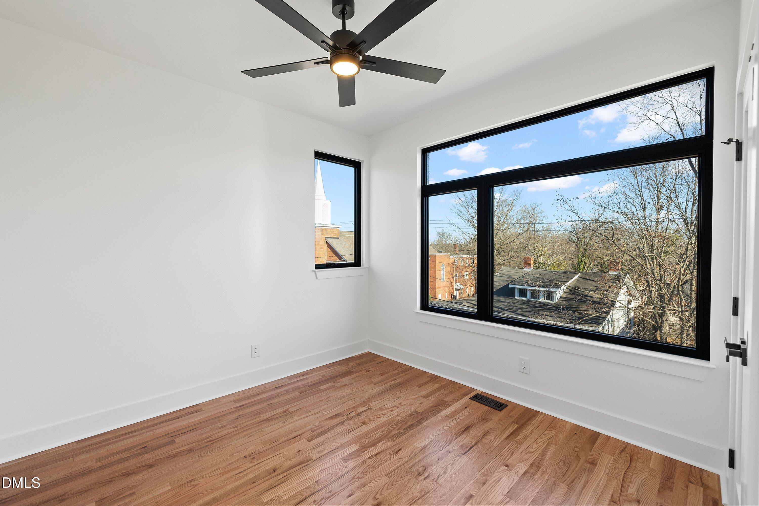 Empty room with hardwood floors, a ceiling fan, and large windows overlooking trees and nearby buildings.