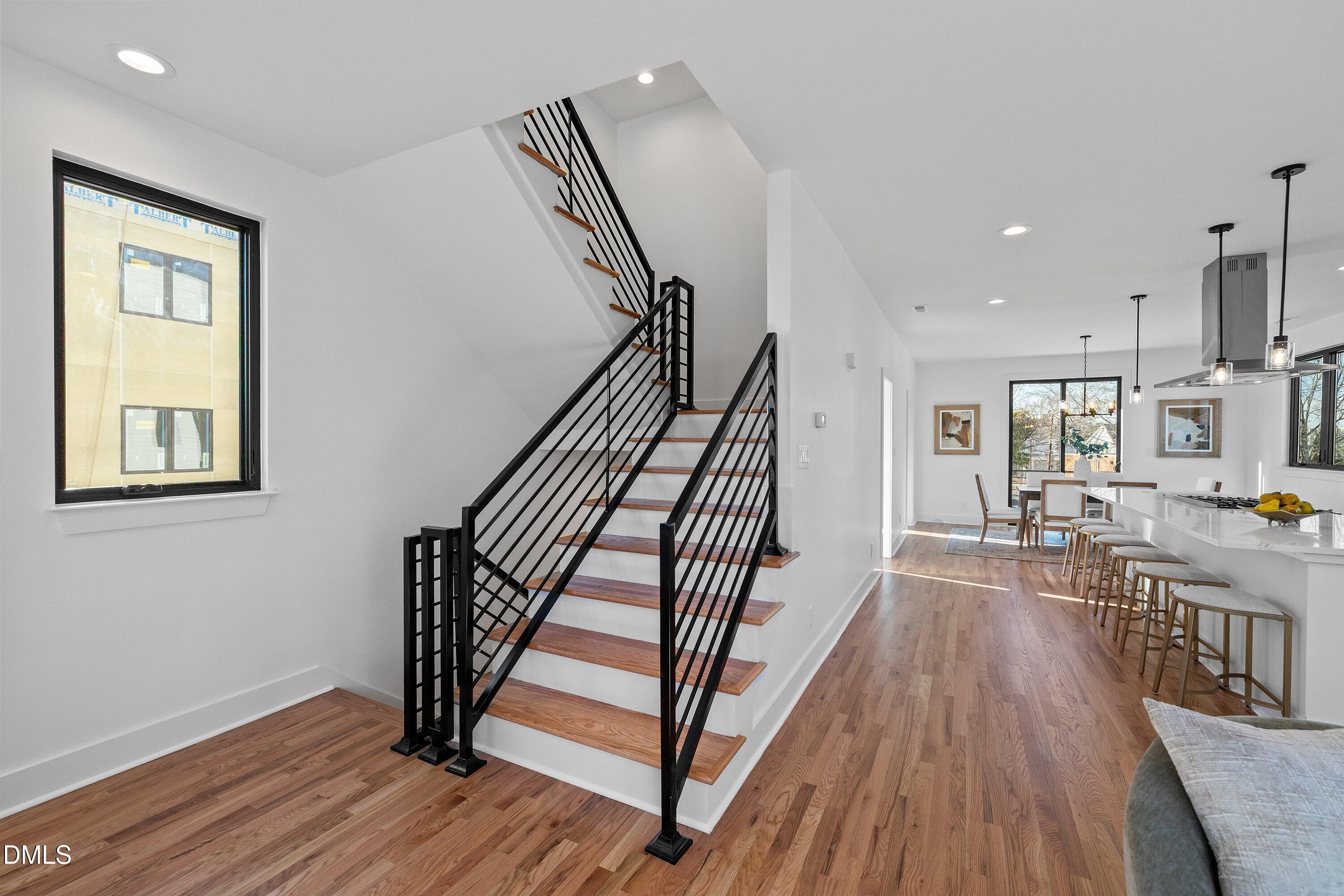 Open-plan interior with a wood-tread staircase, black railings, hardwood floors, and a dining area beside the kitchen.