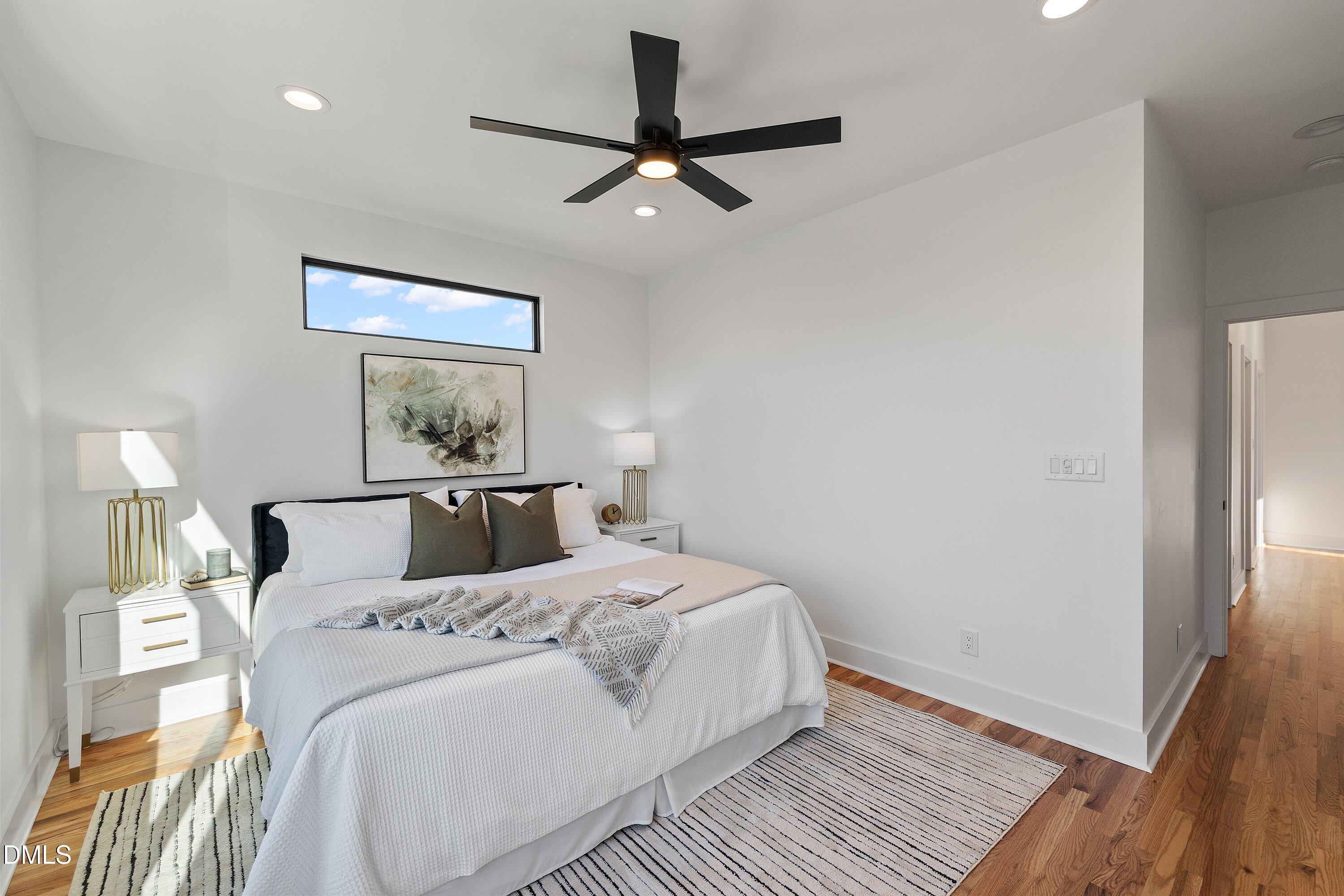 Bright bedroom with a white bed, black ceiling fan, high window, and hardwood floors.