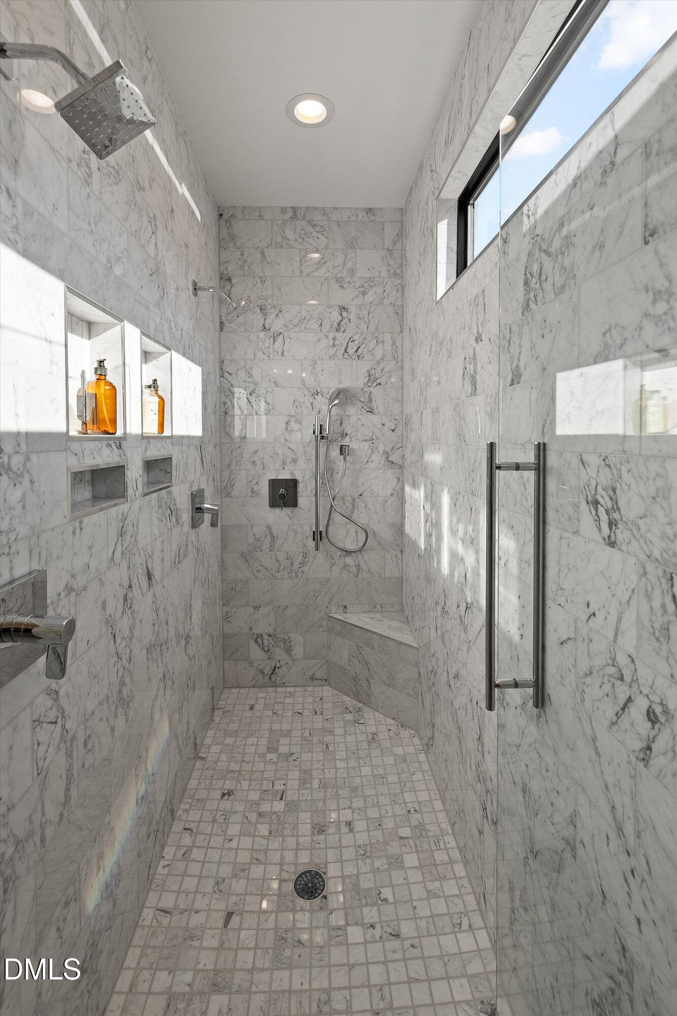 Marble walk-in shower with built-in niches, chrome fixtures, glass door, and high window.