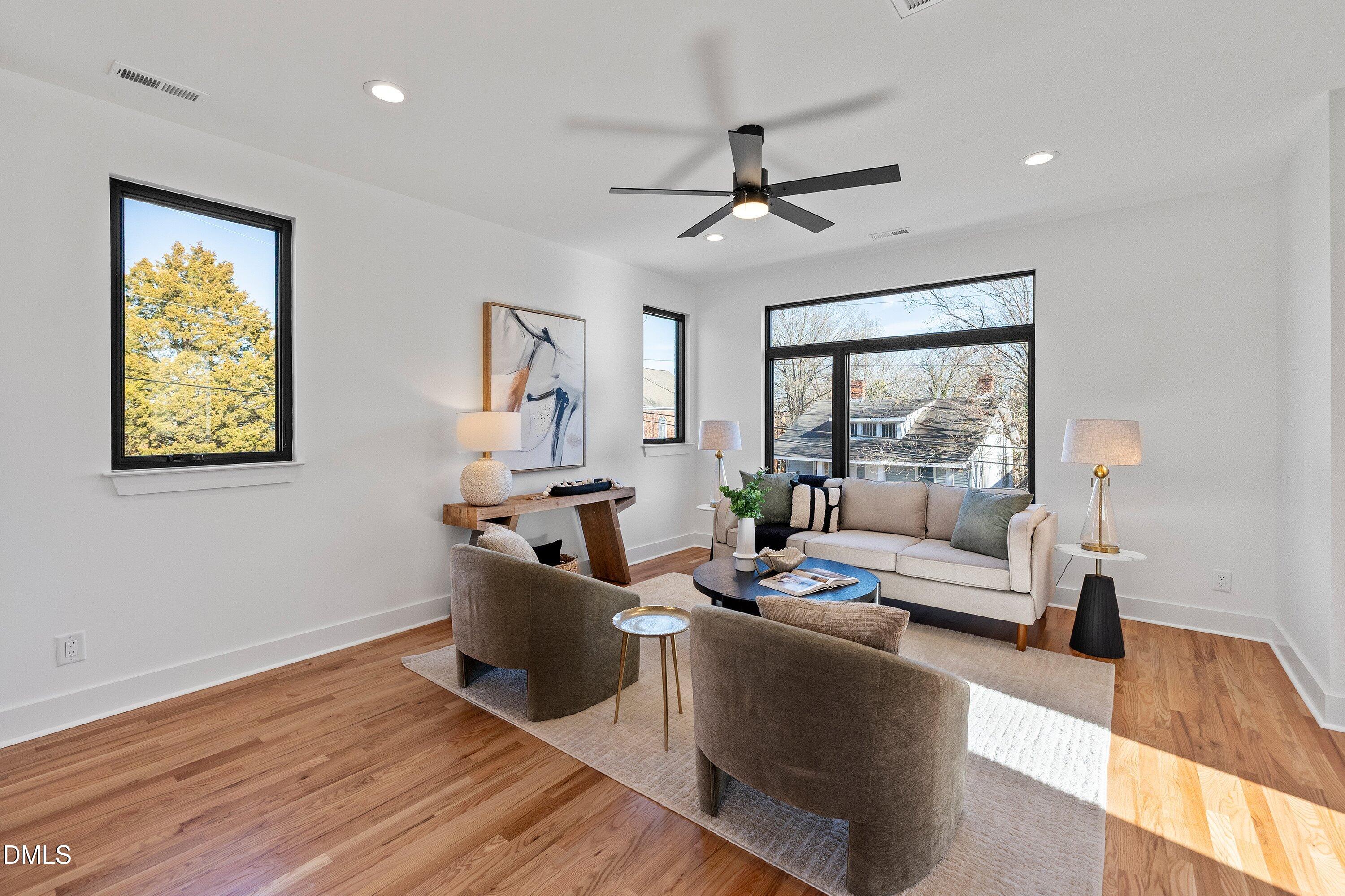 Furnished living room with hardwood floors, large black-framed windows, and a seating area on a pale rug.