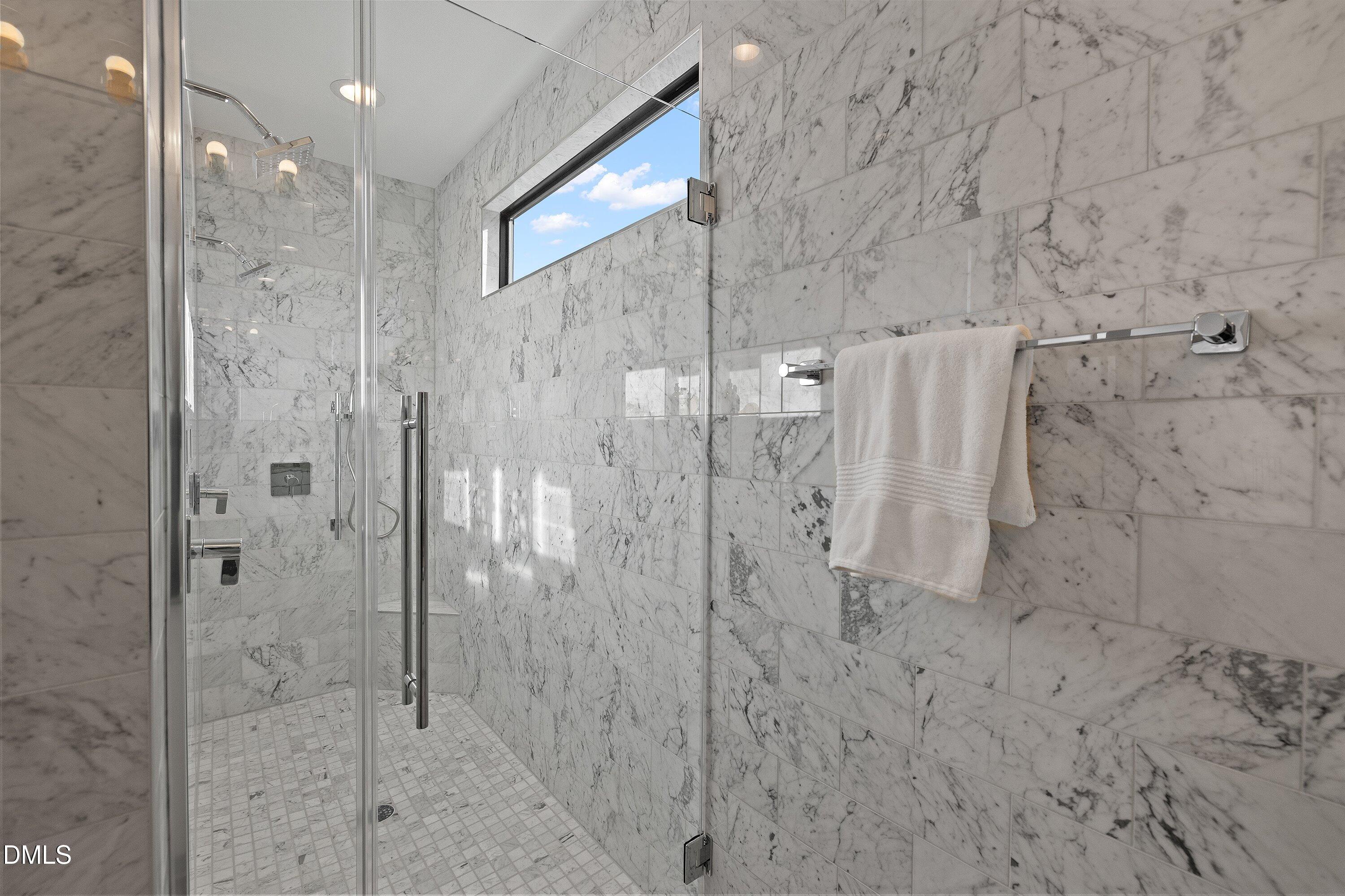 Marble-tiled walk-in shower with frameless glass, chrome fixtures, a high window, and a white towel.