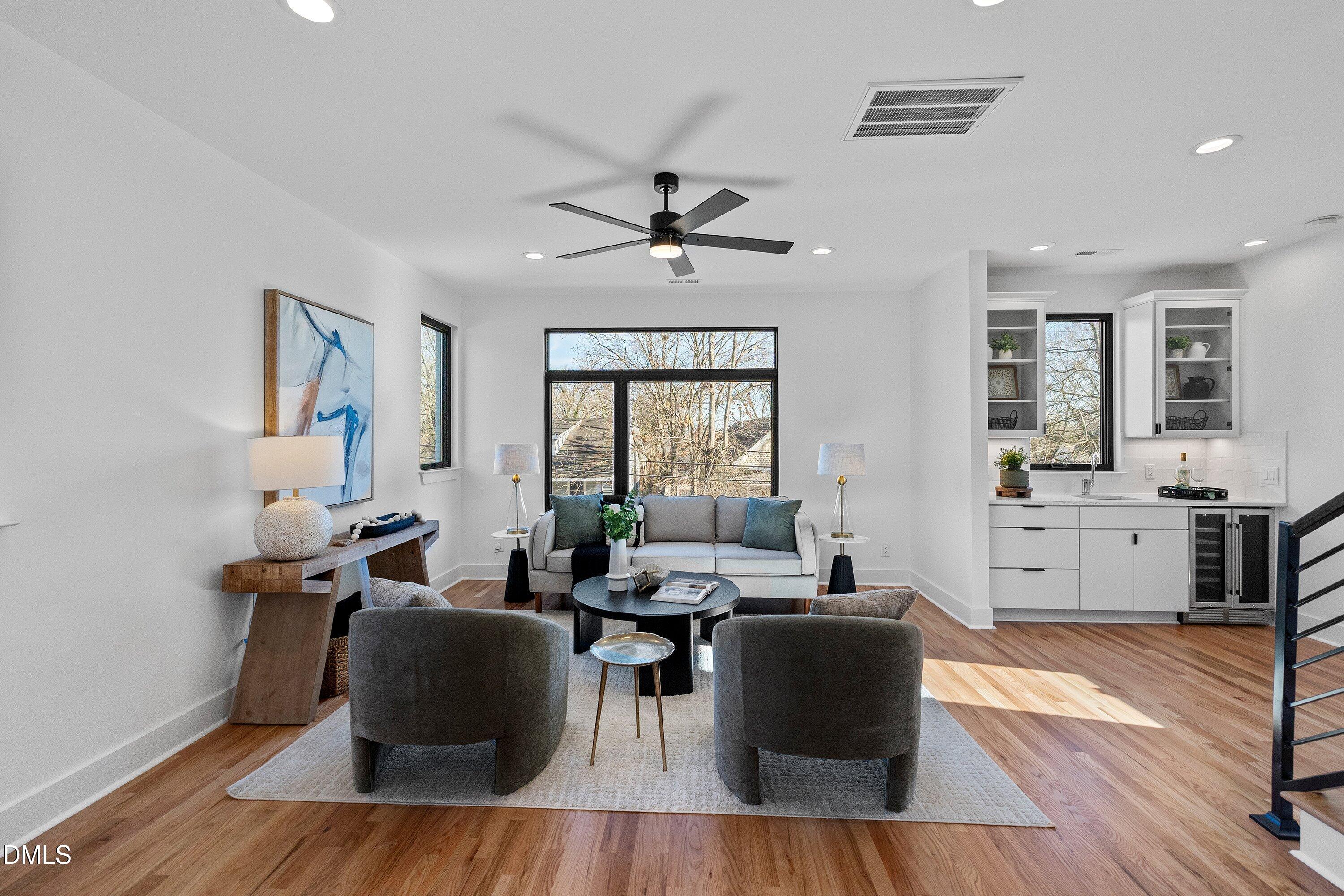 Staged living room with hardwood floors, large windows, and an adjoining modern kitchen.