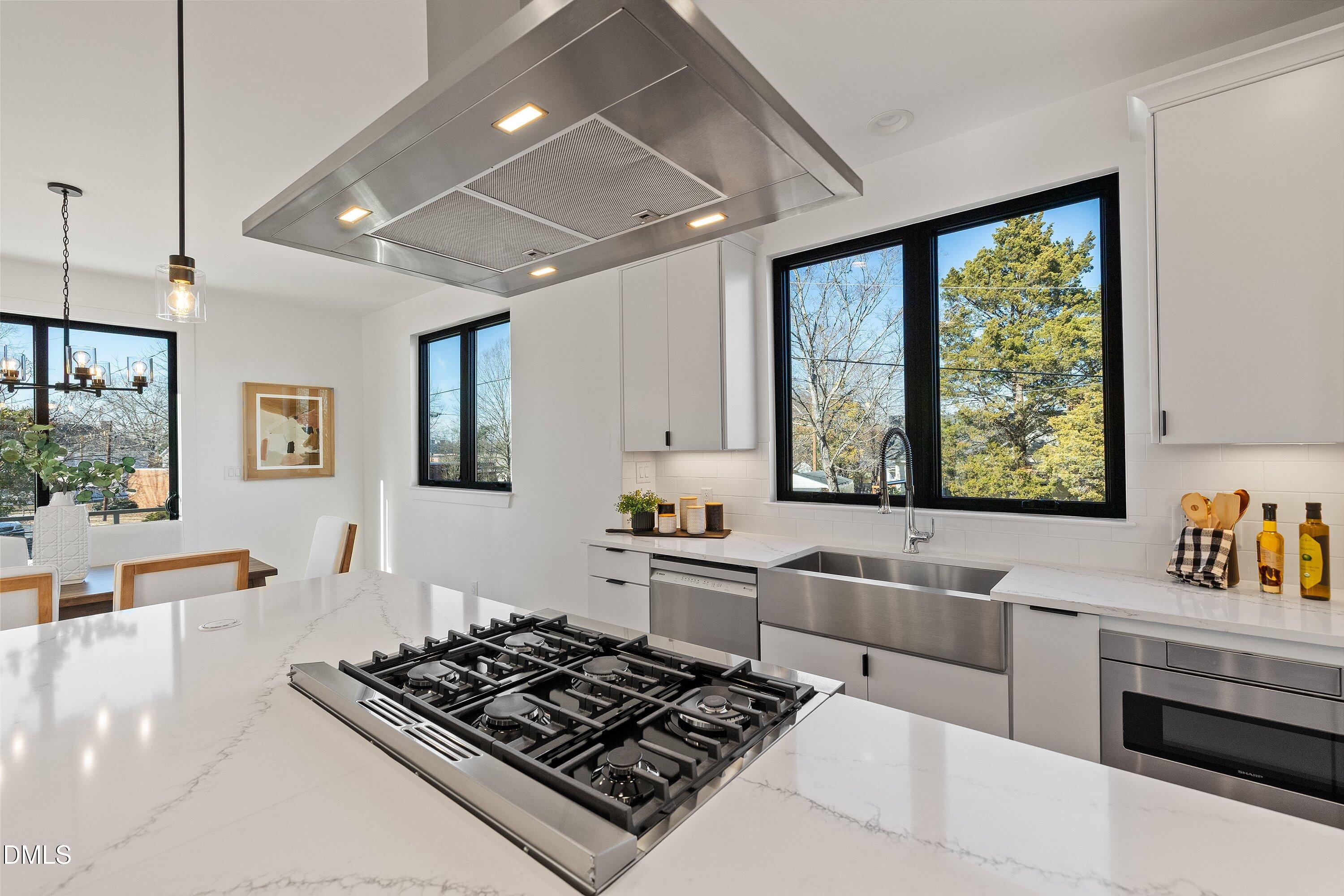 Modern white kitchen with a large cooktop island, stainless-steel hood, and windows overlooking trees.