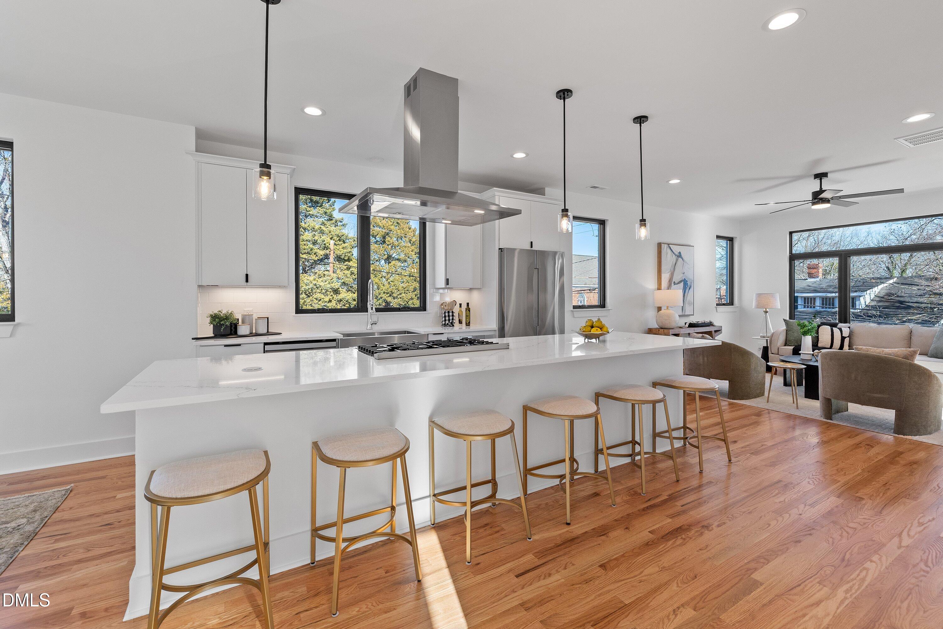 Open-plan kitchen with a long white island, gold-framed stools, stainless steel appliances, and hardwood floors.