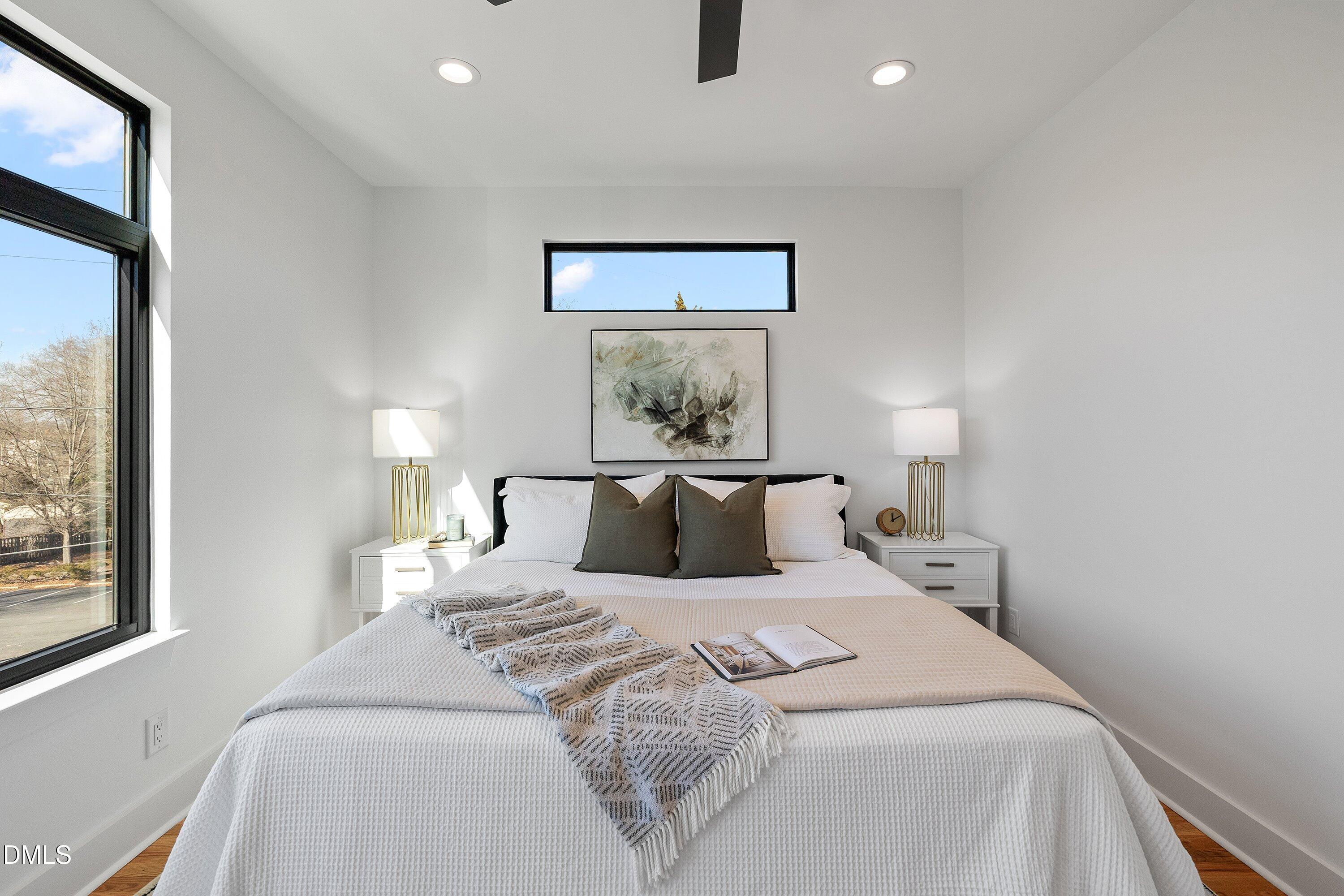 Staged bedroom with a white bed, dark pillows, abstract artwork, and tall black-framed windows.