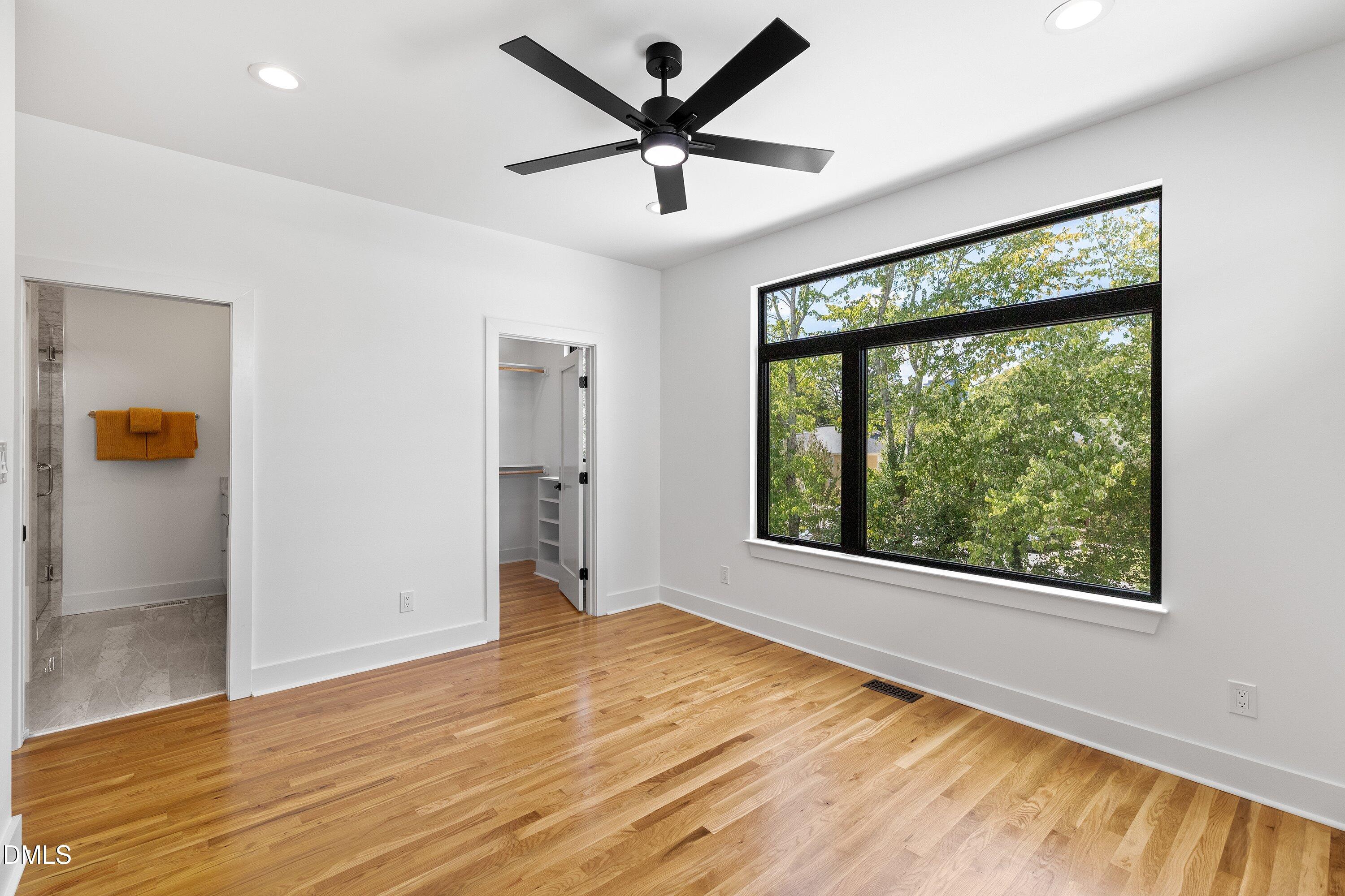 Empty bedroom with hardwood floors, a large window overlooking trees, and an adjoining bathroom and closet.