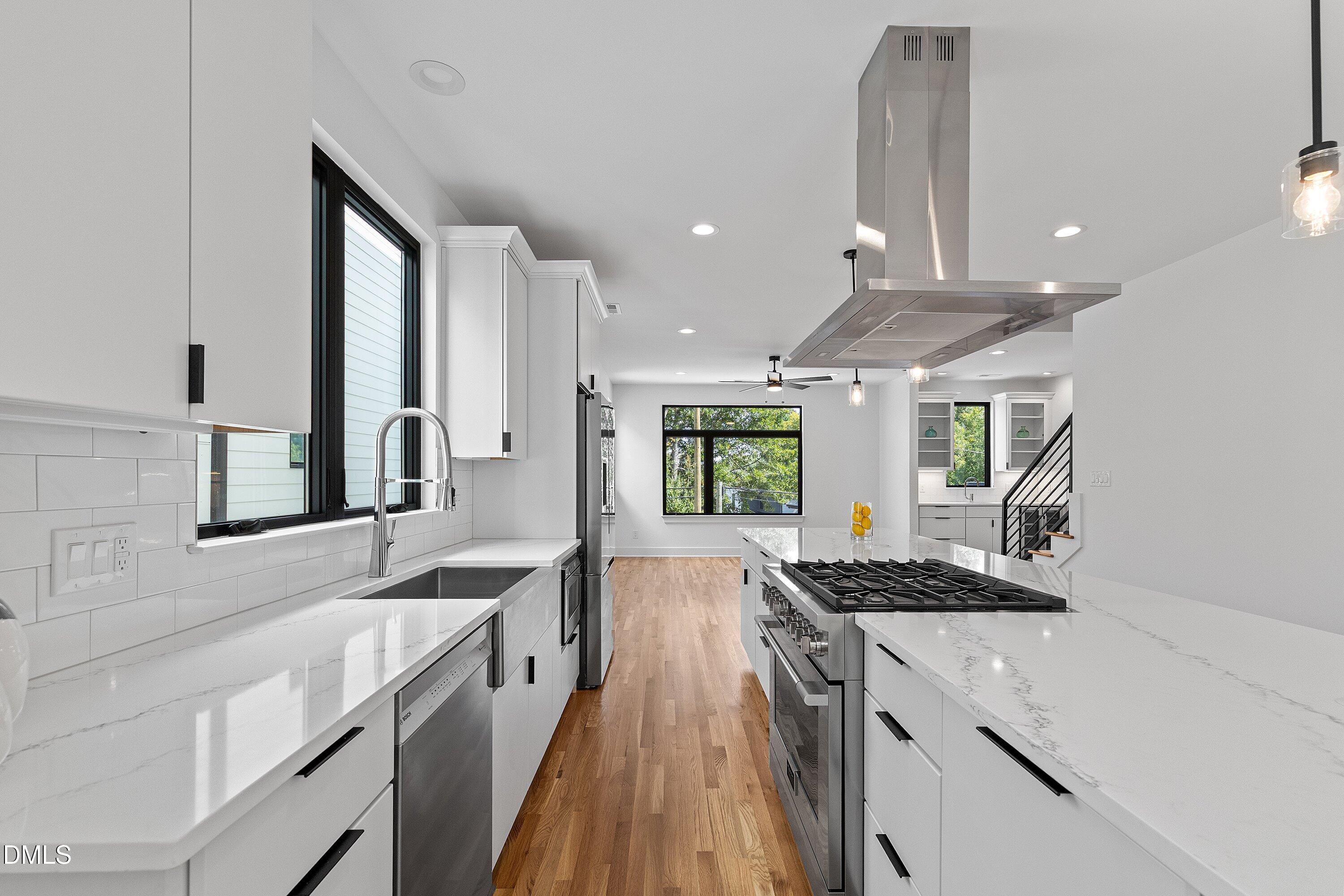 Modern white kitchen with a large cooktop island, stainless-steel hood, and warm wood floors.