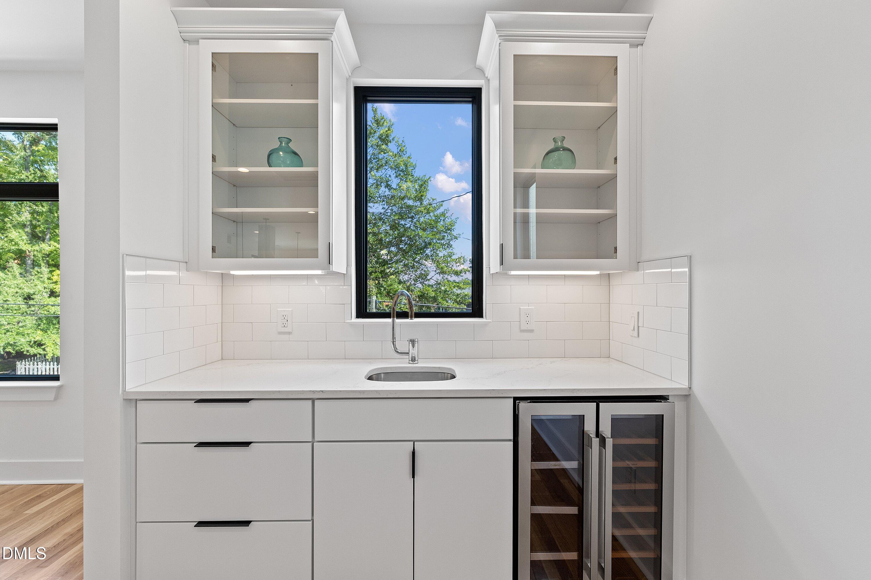 Modern white kitchen with glass-front cabinets, a window above the sink, and a dual-zone wine refrigerator.