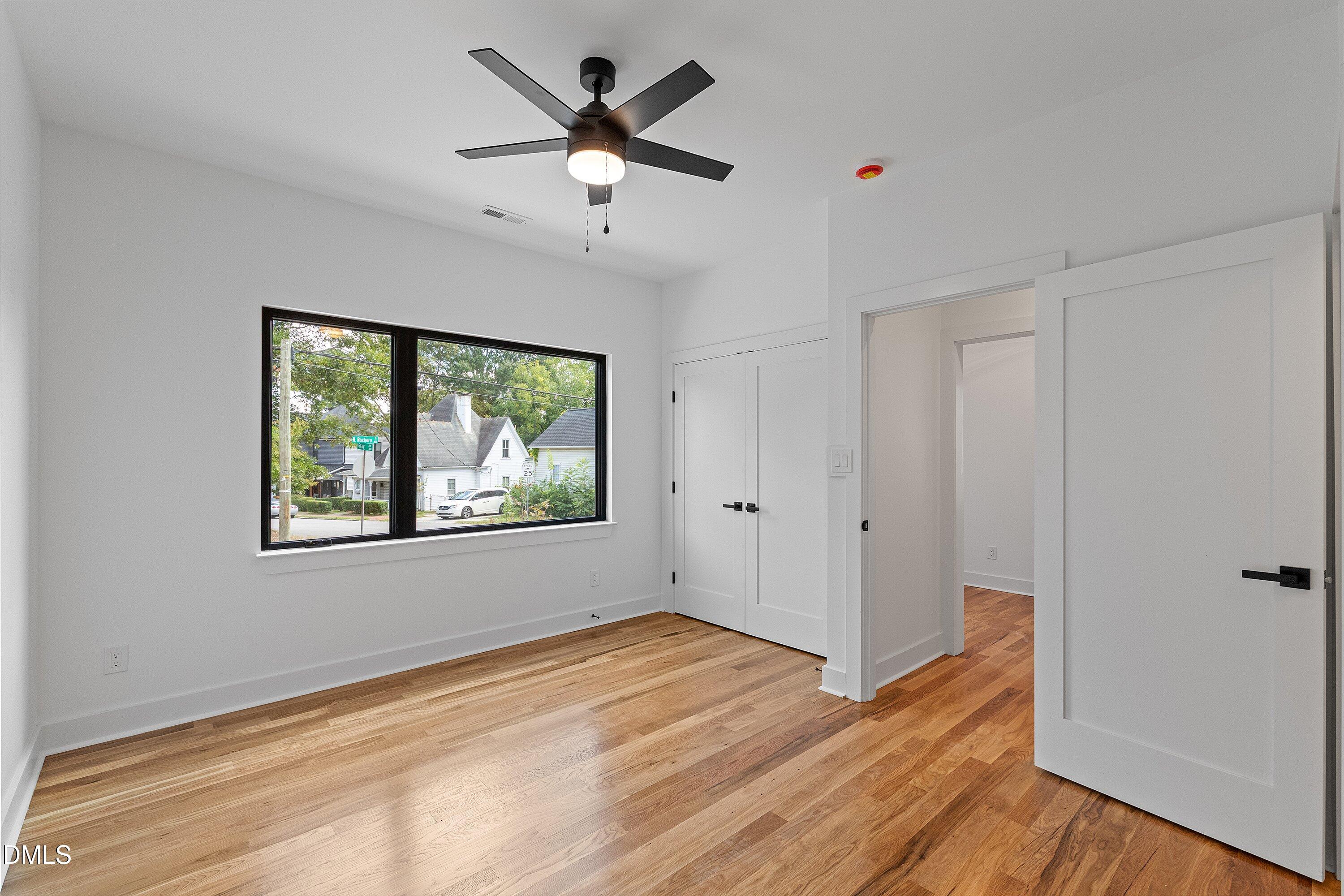 Vacant bedroom with hardwood floors, a black ceiling fan, and a large window overlooking the street.