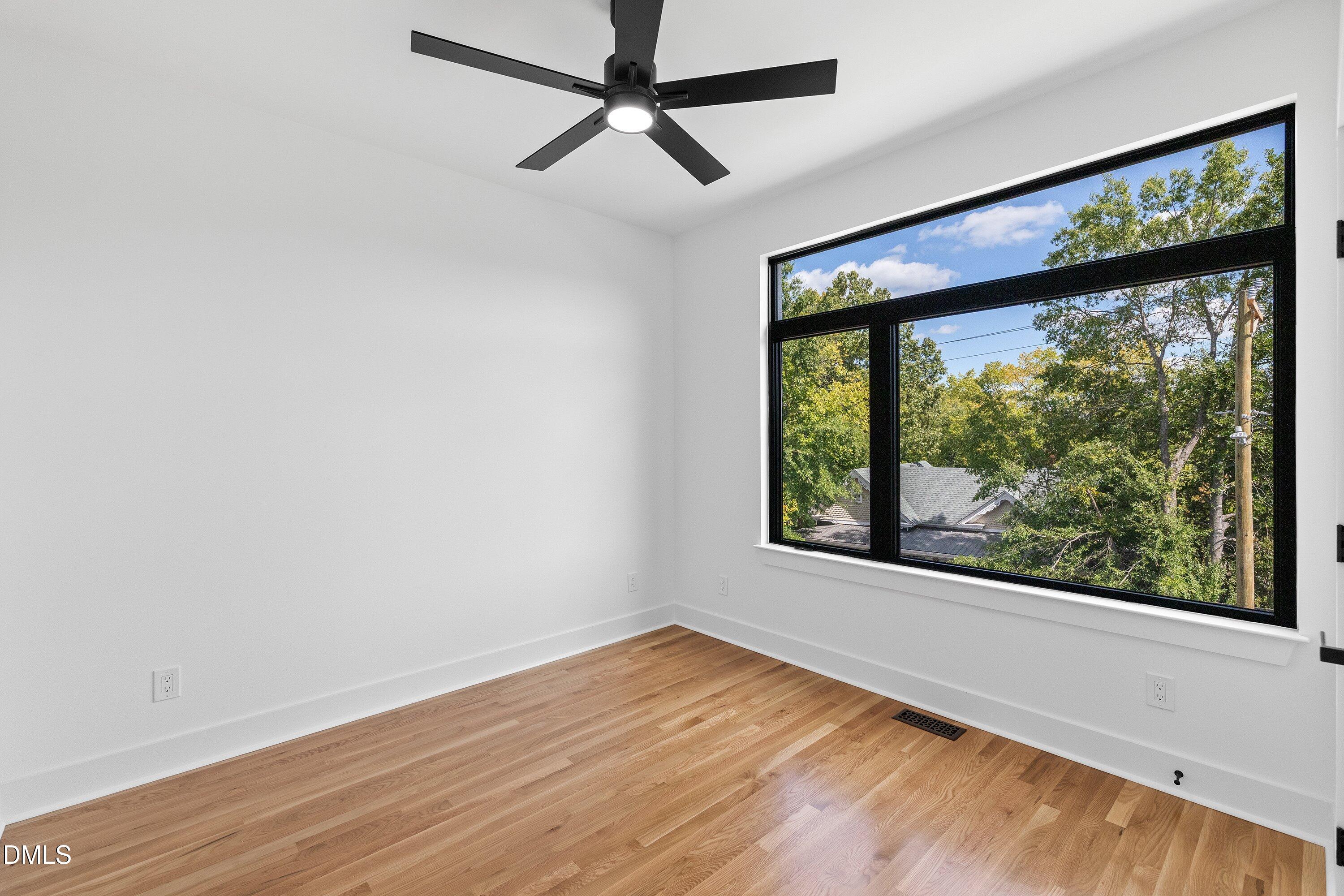 Vacant room with hardwood floors, a black ceiling fan, and a large window overlooking leafy trees.
