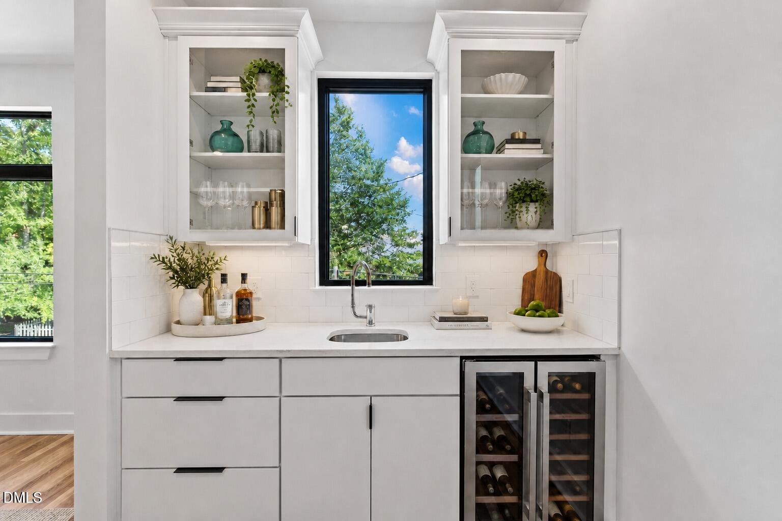 White beverage station with a sink, glass-front cabinets, and twin wine refrigerators beneath a tall window.