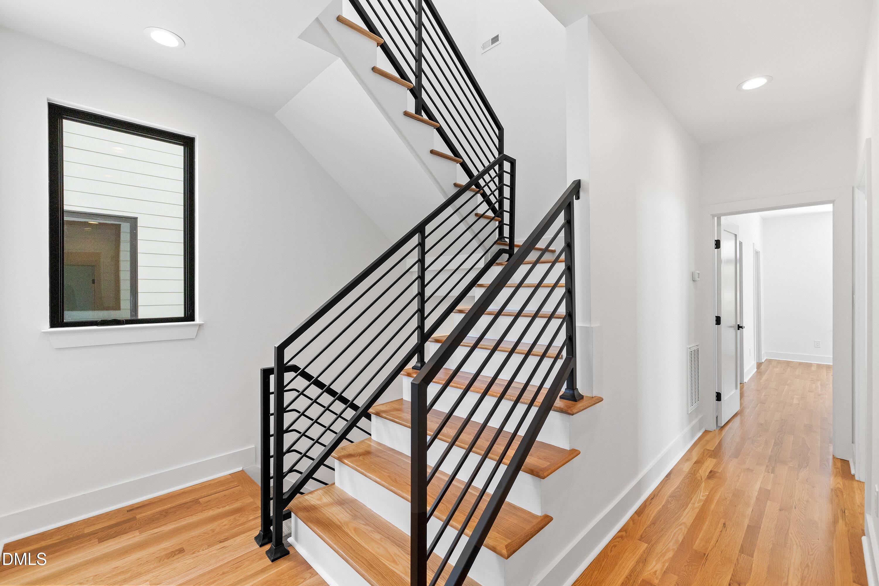 Wood-tread staircase with black metal railings beside a bright hardwood-floored hallway.