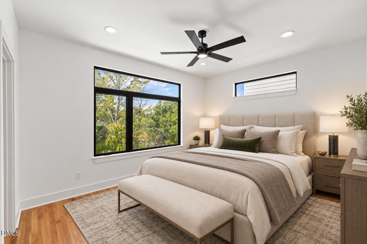 Staged bedroom with a large bed, black-framed windows overlooking trees, and a black ceiling fan.