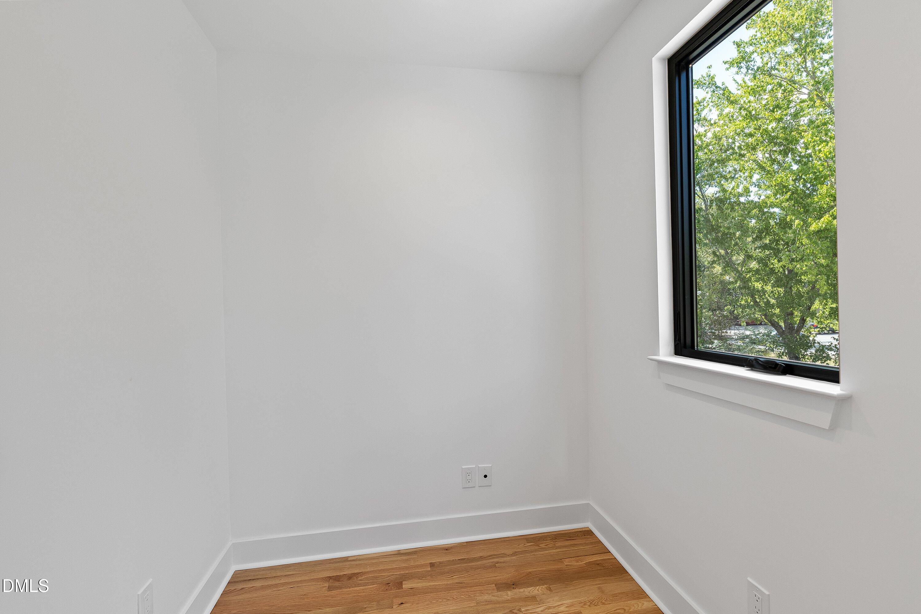Vacant room with hardwood flooring and a large black-framed window overlooking trees.