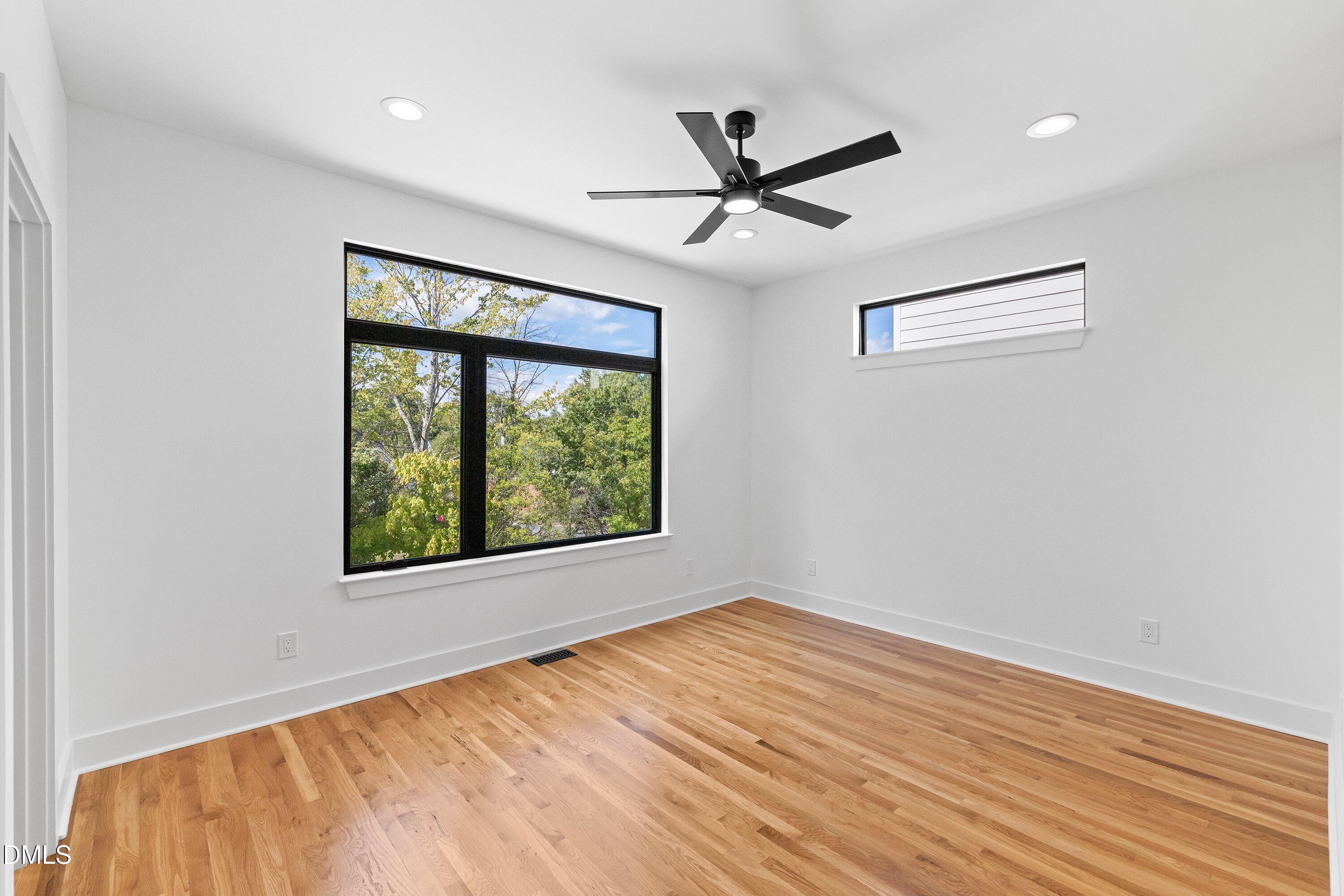 Bright vacant room with hardwood floors, large black-framed windows, and a modern ceiling fan.