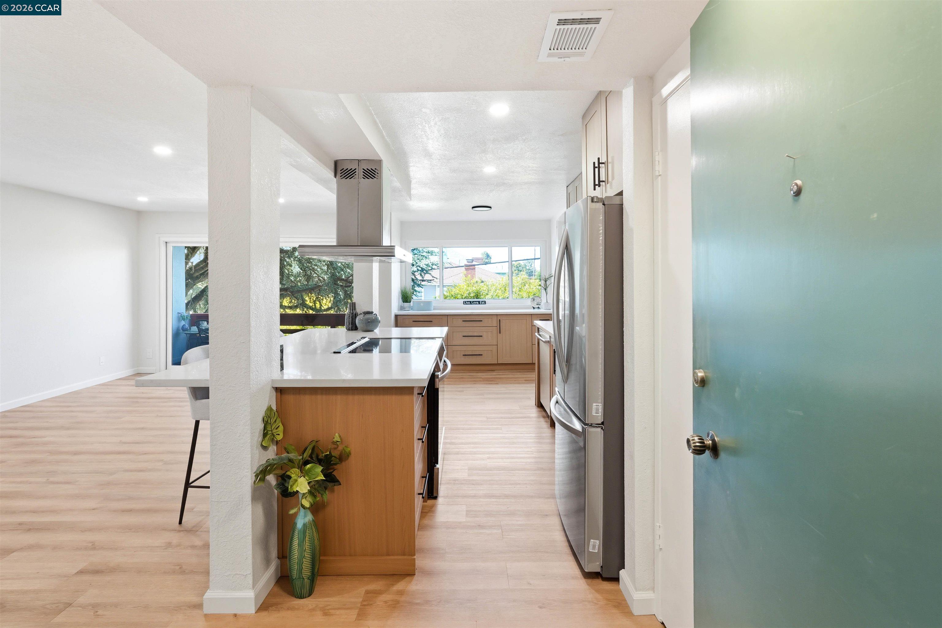 Open-plan kitchen with a white cooktop island, wood cabinetry, stainless-steel appliances, and a teal entry door.