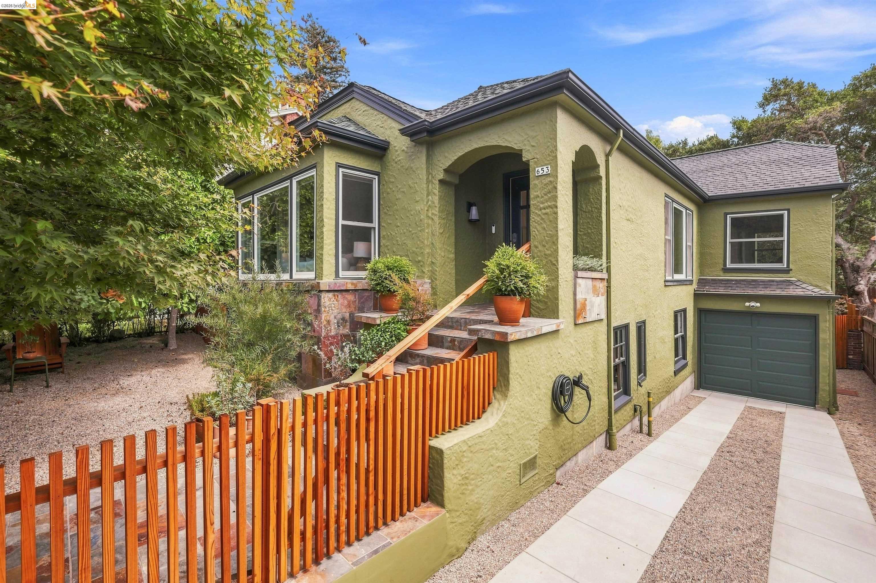Sage-green stucco home with an arched entry, tiled steps, wooden railing, and attached garage.