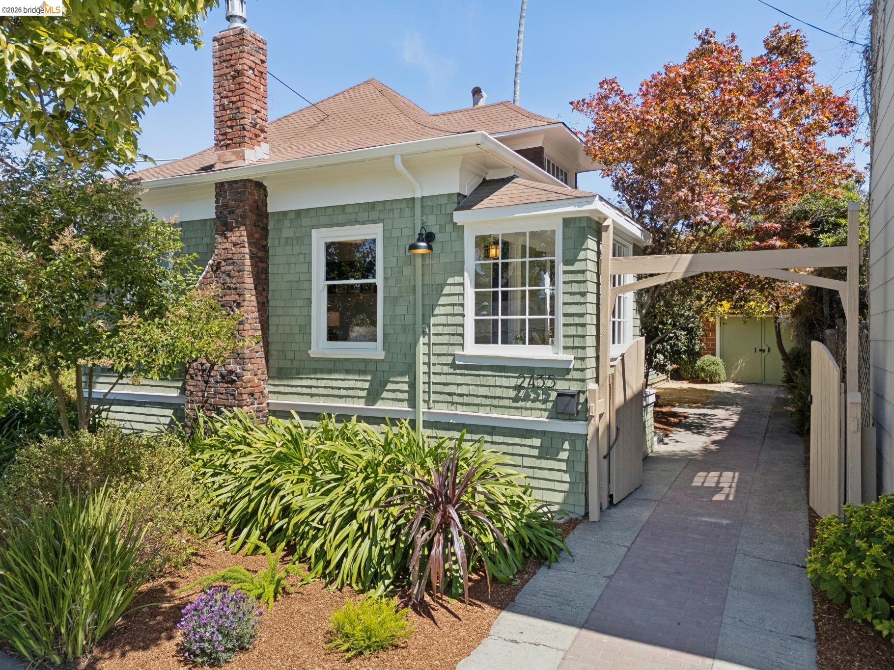 Green shingle-sided home with a brick chimney, landscaped garden, and paved driveway to a detached garage.