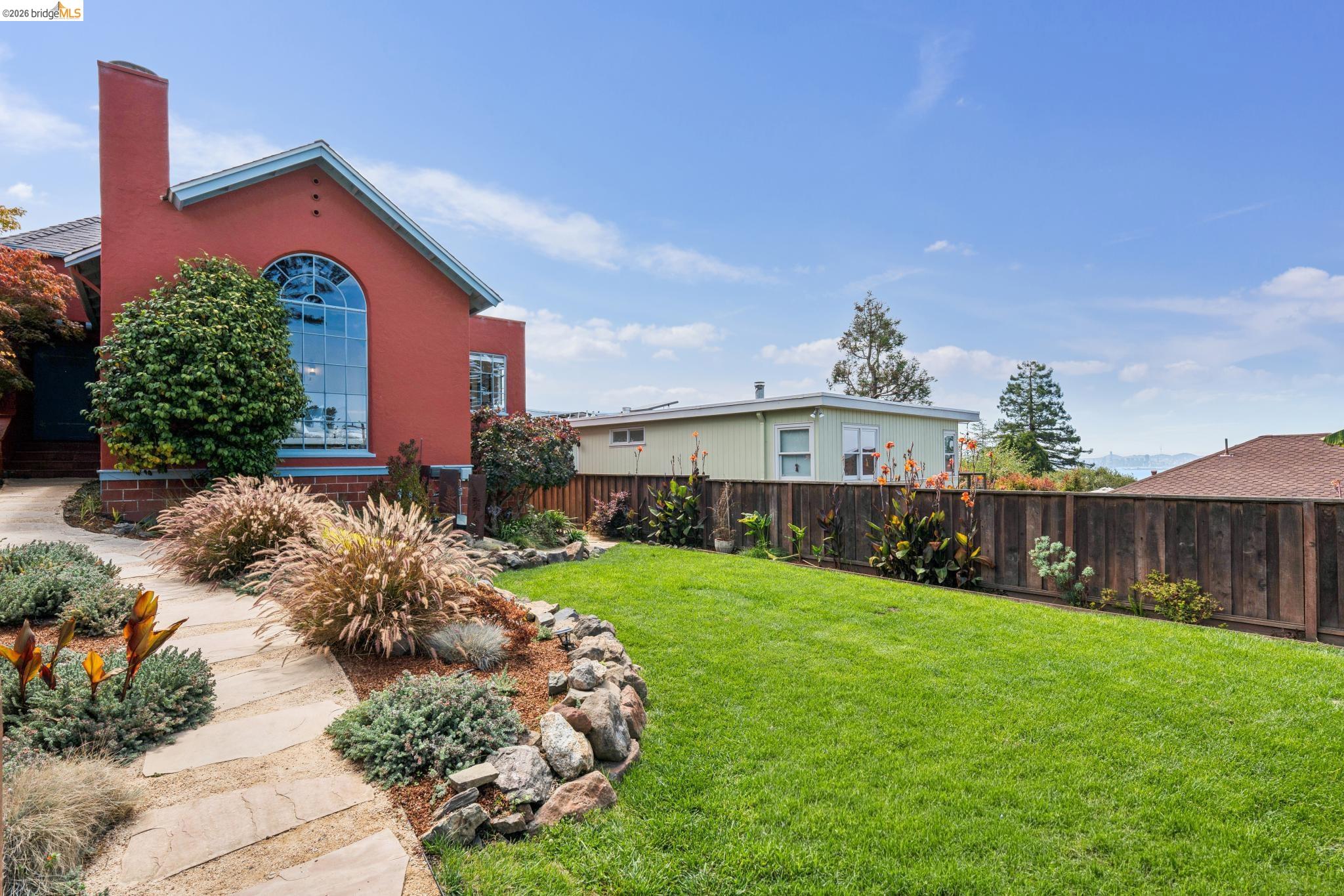 Red contemporary house with an arched window, landscaped stone path, and fenced green lawn.
