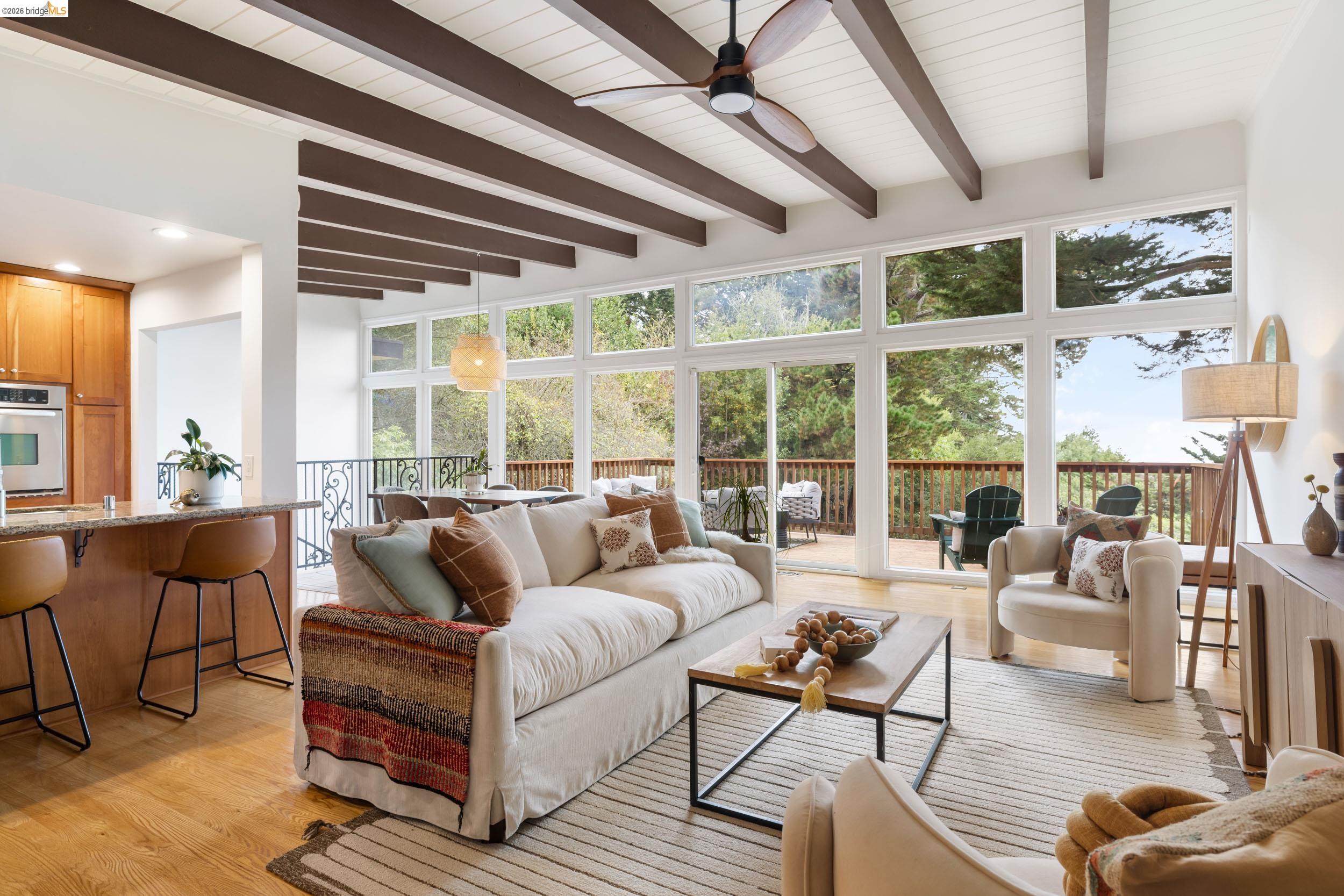 Living room with exposed ceiling beams, a wall of windows, and a deck overlooking trees.