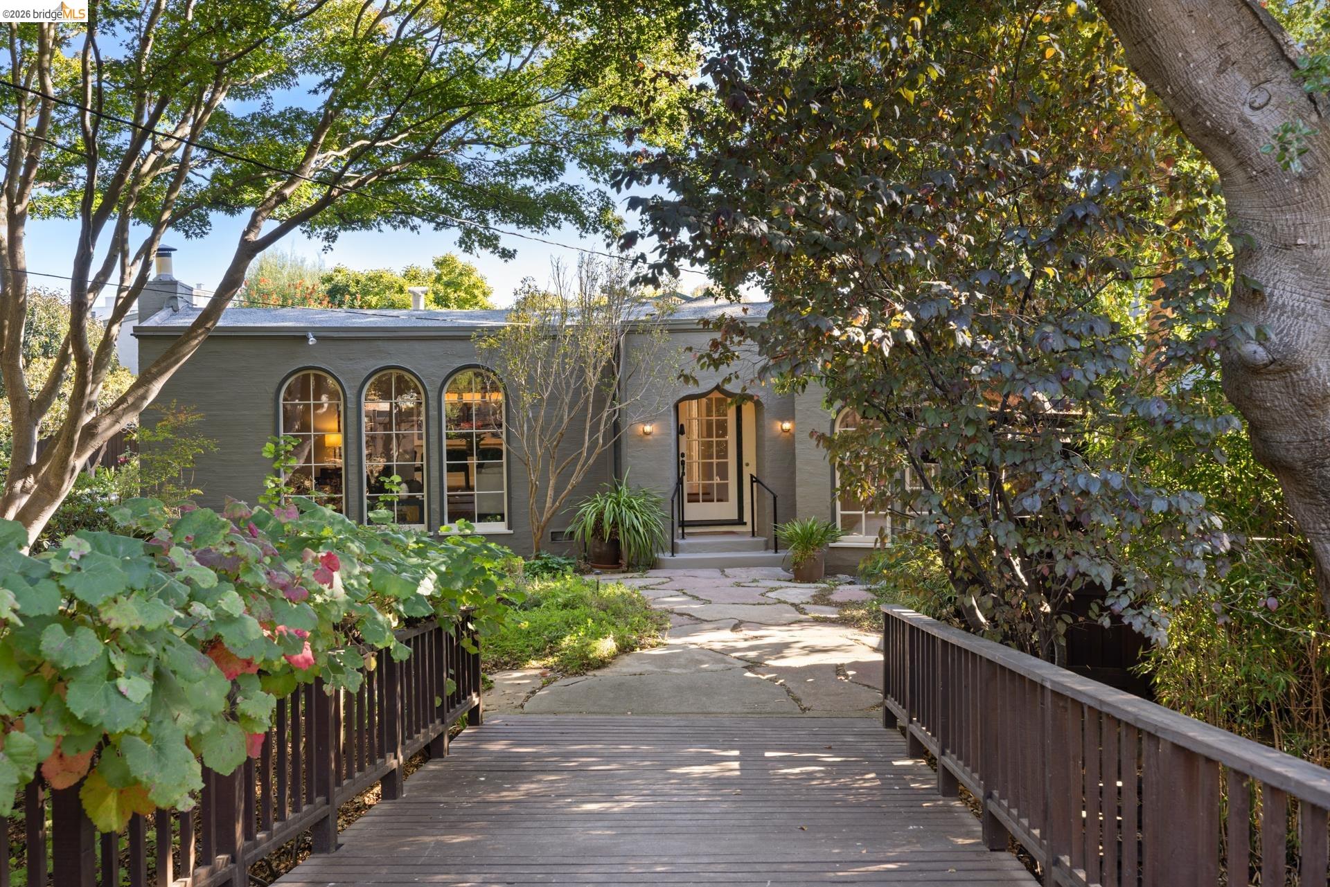 Gray house with arched windows, approached by a wooden footbridge through mature garden landscaping.