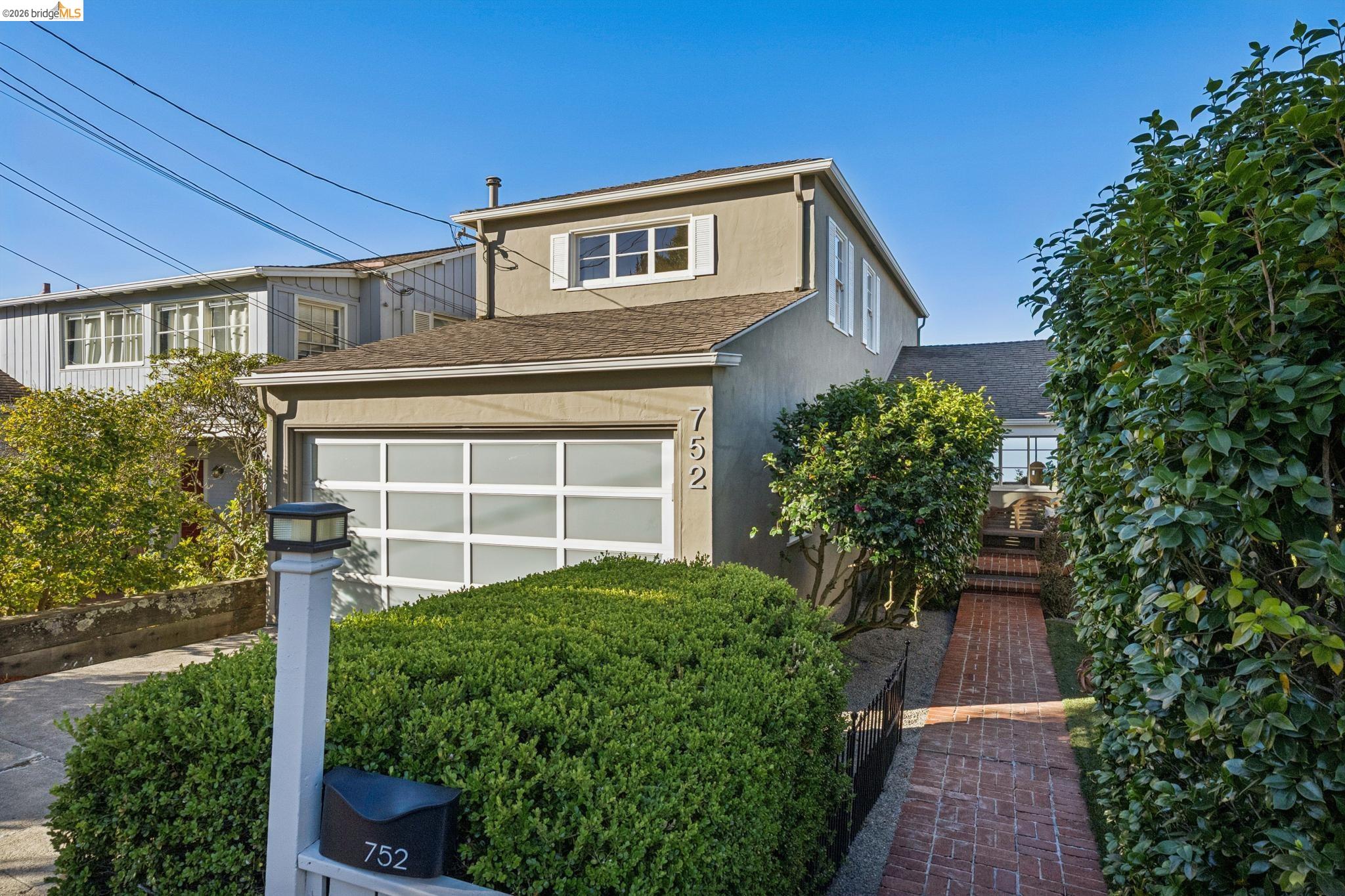 Two-story home with a white garage door, trimmed hedges, and a brick walkway to the entrance.